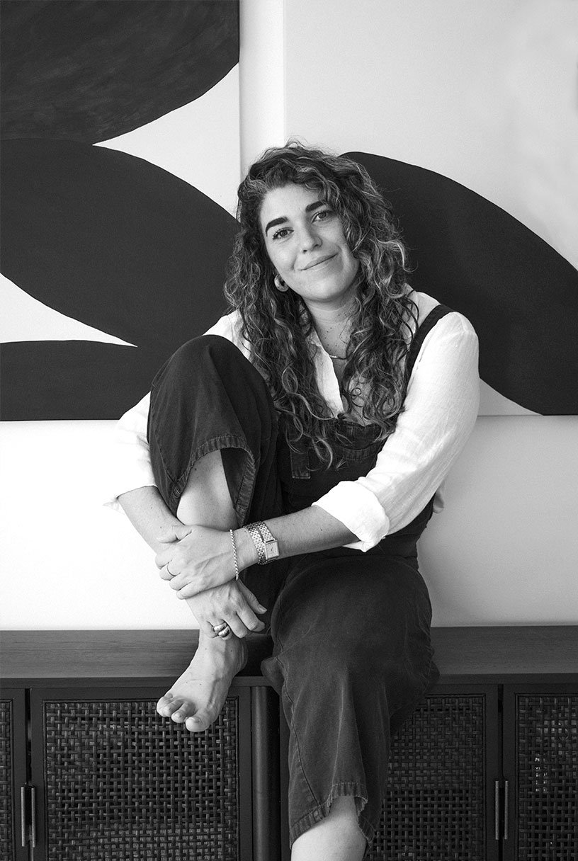 A woman with curly hair sitting on a bench with one leg crossed over the other. She is smiling slightly at the camera and is wearing a light-colored shirt and dark pants. Behind her is a large, abstract, black-and-white painting.