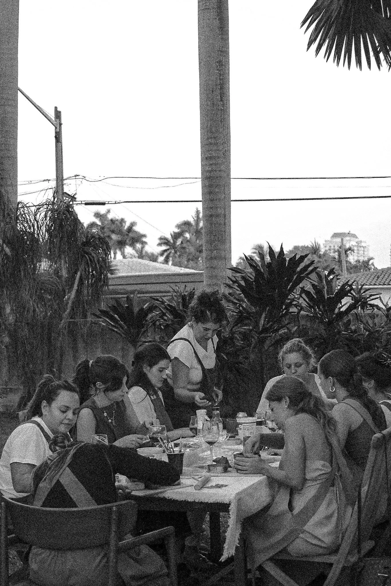 Group of women sitting around an outdoor table, engaged in a craft or activity, with greenery and utility poles in the background.