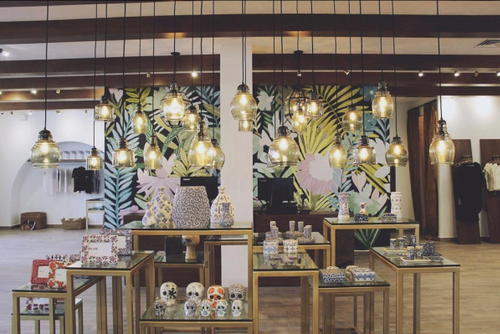 Display of decorative skulls and small items on tables in front of a colorful tropical wallpaper, with hanging light fixtures overhead.