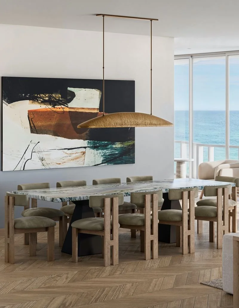 Modern dining room with a marble table, eight beige cushioned chairs, large abstract art painting, a golden pendant light, and a view of the ocean through large windows.