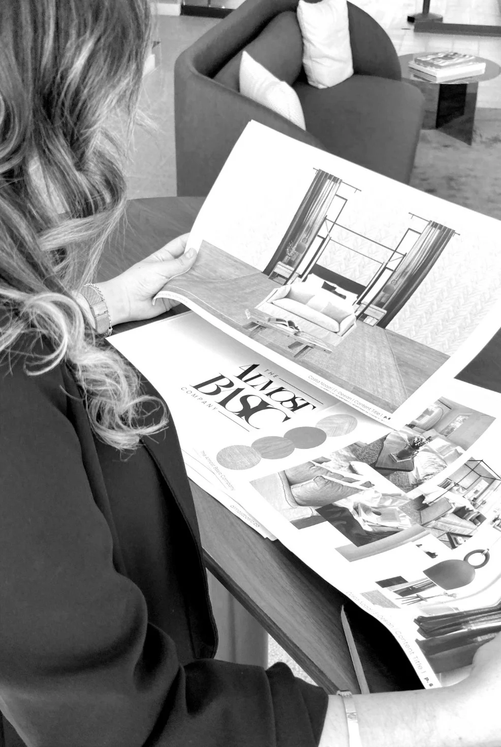 A woman sitting at a table reviewing a furniture catalog, with a living room area in the background.