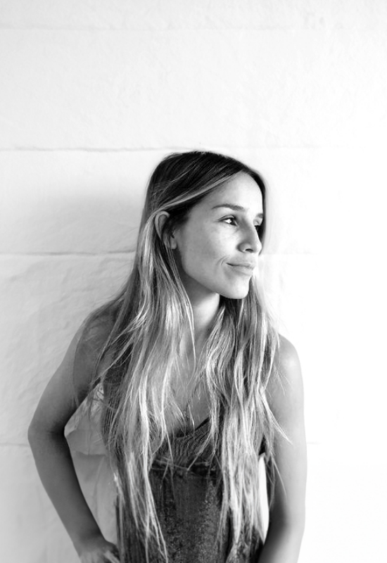 A young woman with long, wavy hair looking to her right, standing against a plain wall.
