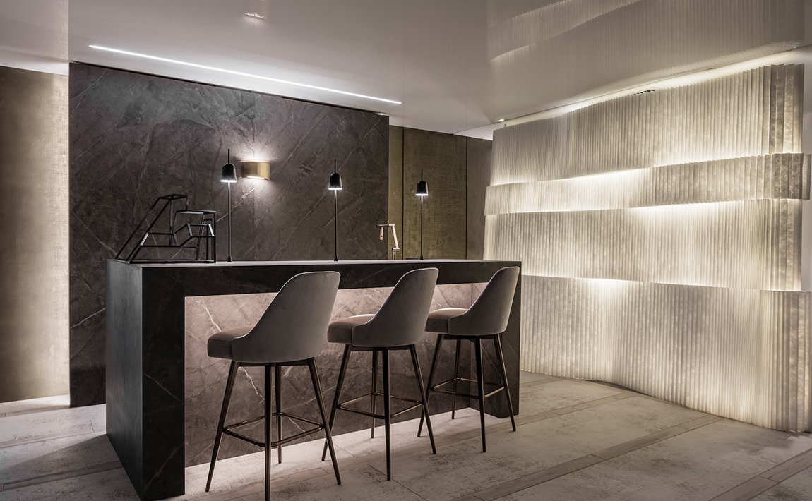Modern interior space with dark marble bar, three gray bar stools, backlit textured wall, and minimalist lighting.