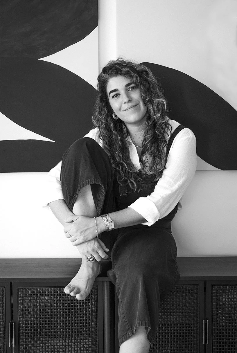 A young woman with curly hair, wearing a white shirt and dark overalls, sitting on a cabinet with her leg crossed over the other, smiling with a closed-mouth smile, in front of an abstract black and white painting.