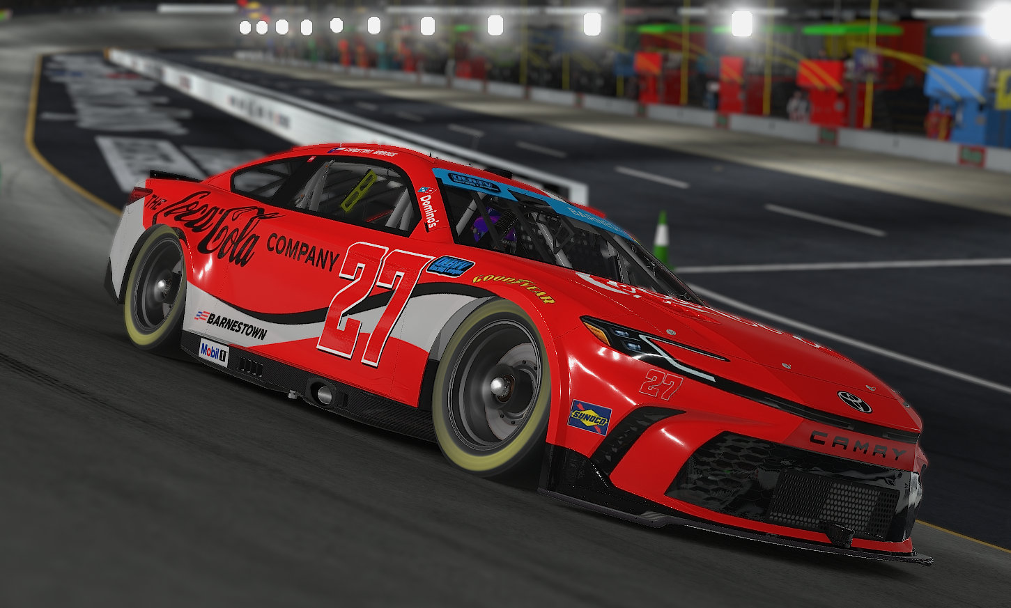 Red Toyota Camry race car with number 27 driving on a race track, surrounded by pit area and barriers.