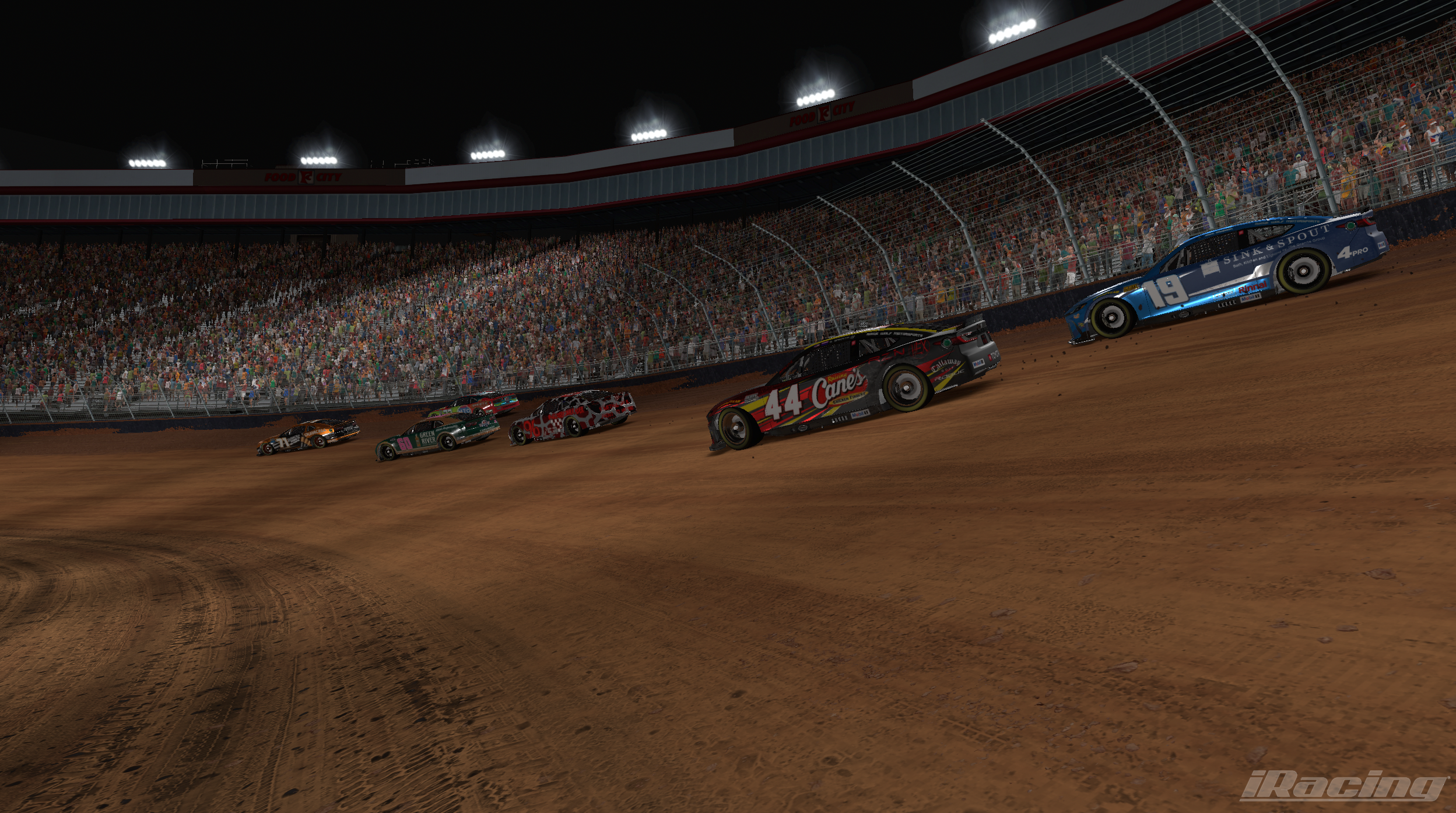 Night race at a dirt track with multiple racing cars, including black, red, green, and blue cars, and a crowd of spectators in the stands.