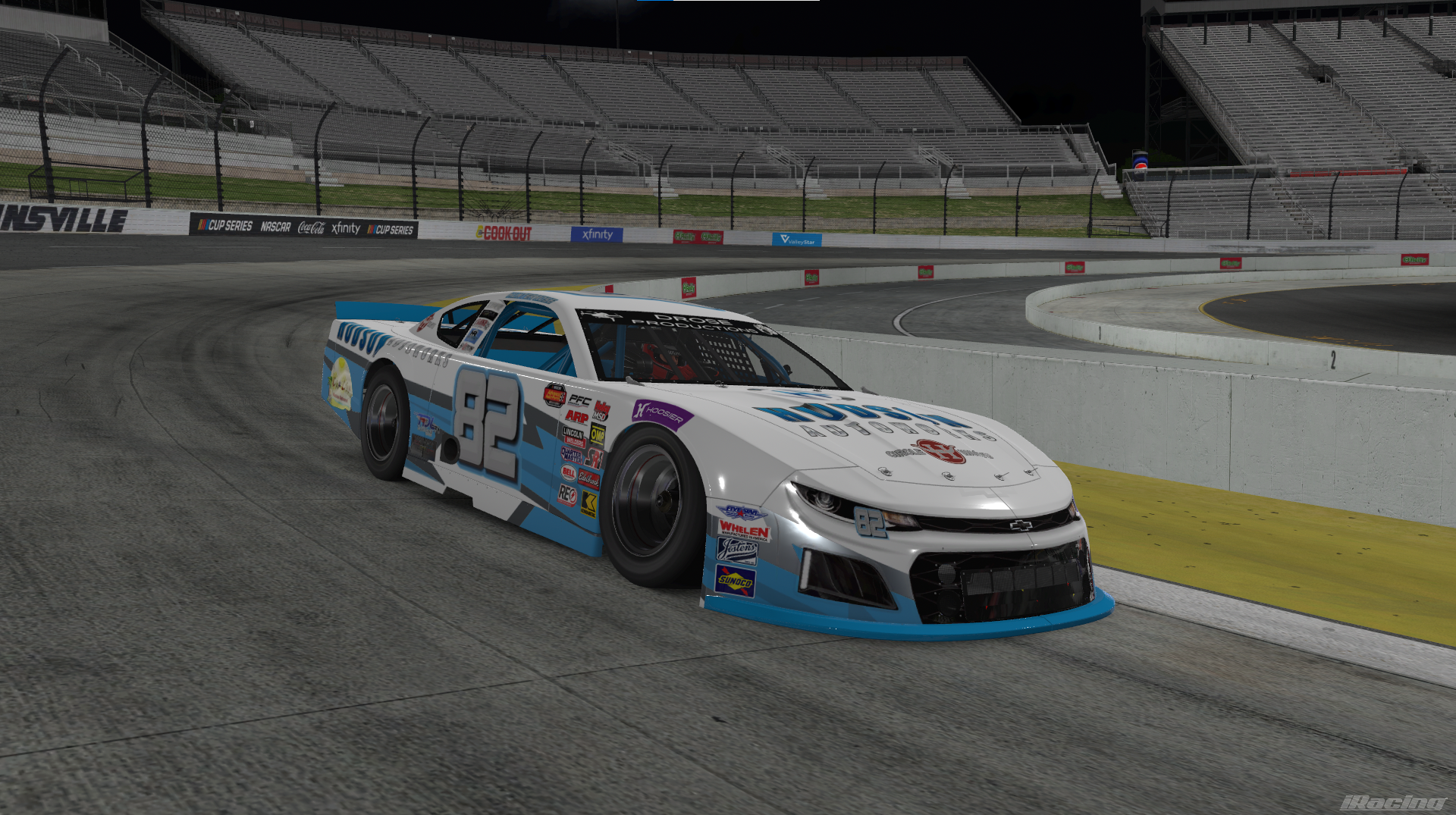 A white and blue race car driven by number 82 speeds around a dirt track at night with empty grandstands and advertising banners in the background.