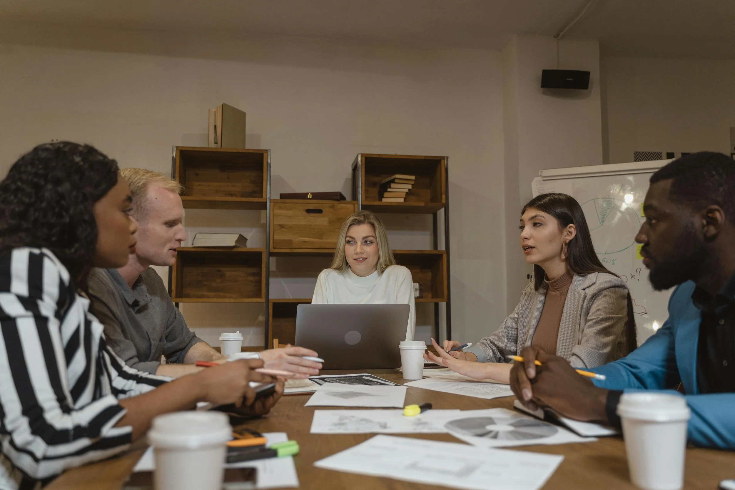 Five diverse people sitting around a wooden table in a meeting room, engaged in discussion, with papers, pens, laptops, and coffee cups in front of them.
