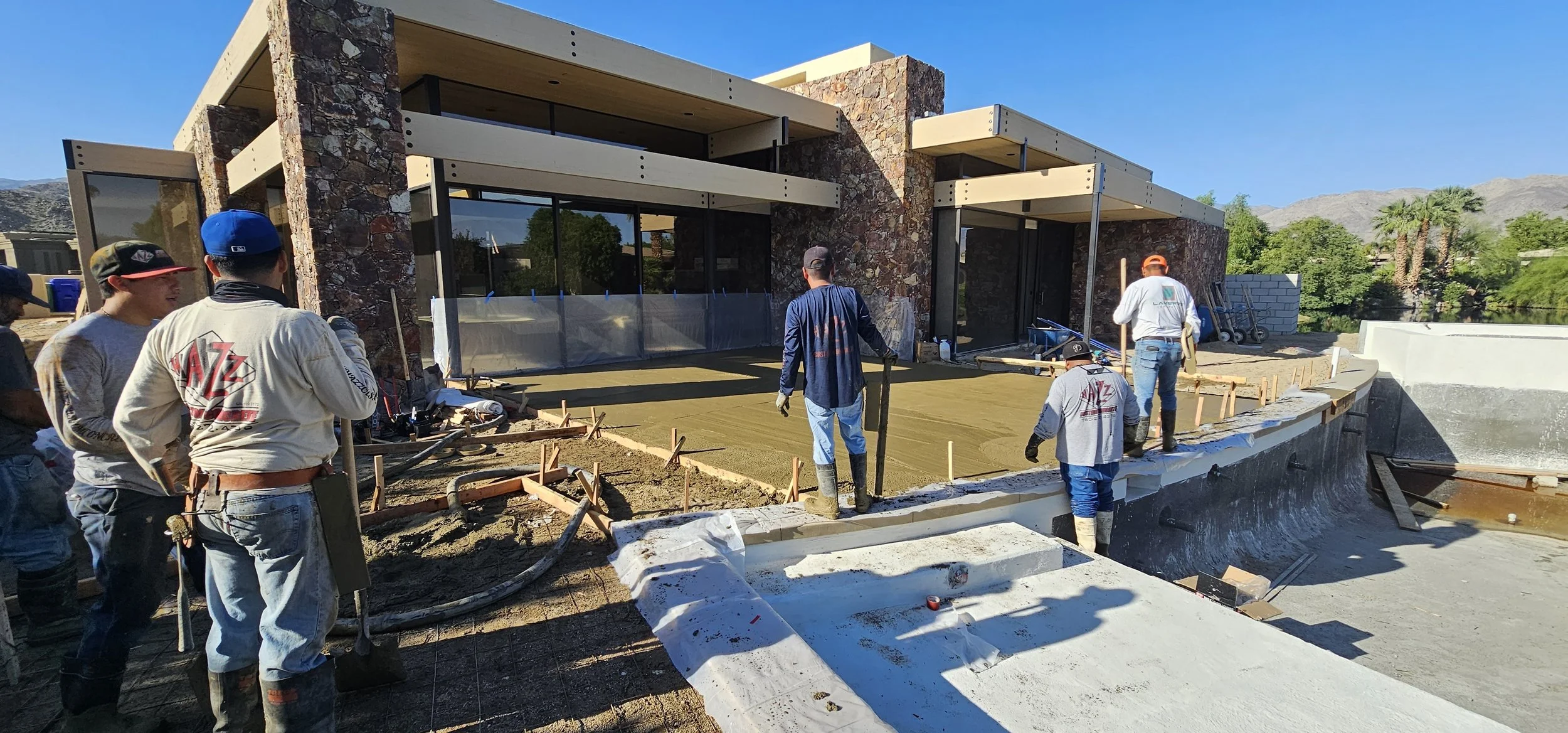 Construction workers pouring concrete on a patio in front of a modern house with stone and glass exterior, surrounded by trees and mountains.