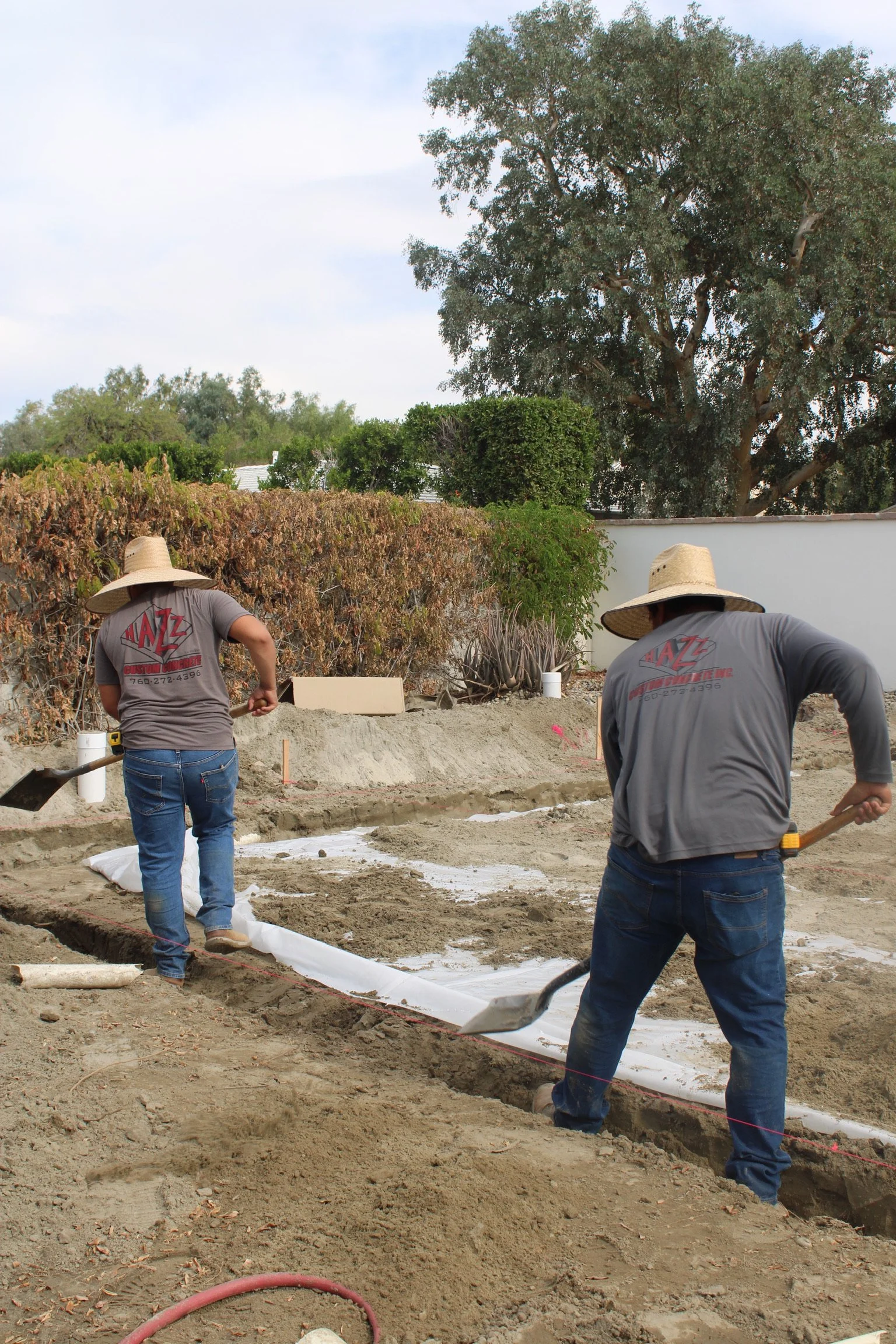 Two workers wearing wide-brimmed hats and gray t-shirts are working on a construction site, installing a white sheet on the ground, with trees and bushes in the background.