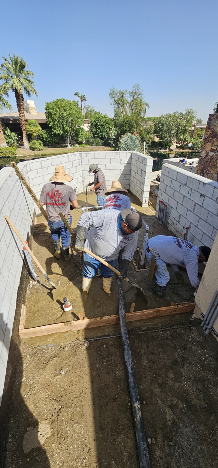 Construction workers laying concrete in a small, partially enclosed backyard with cinder block walls, green trees, and a clear blue sky.
