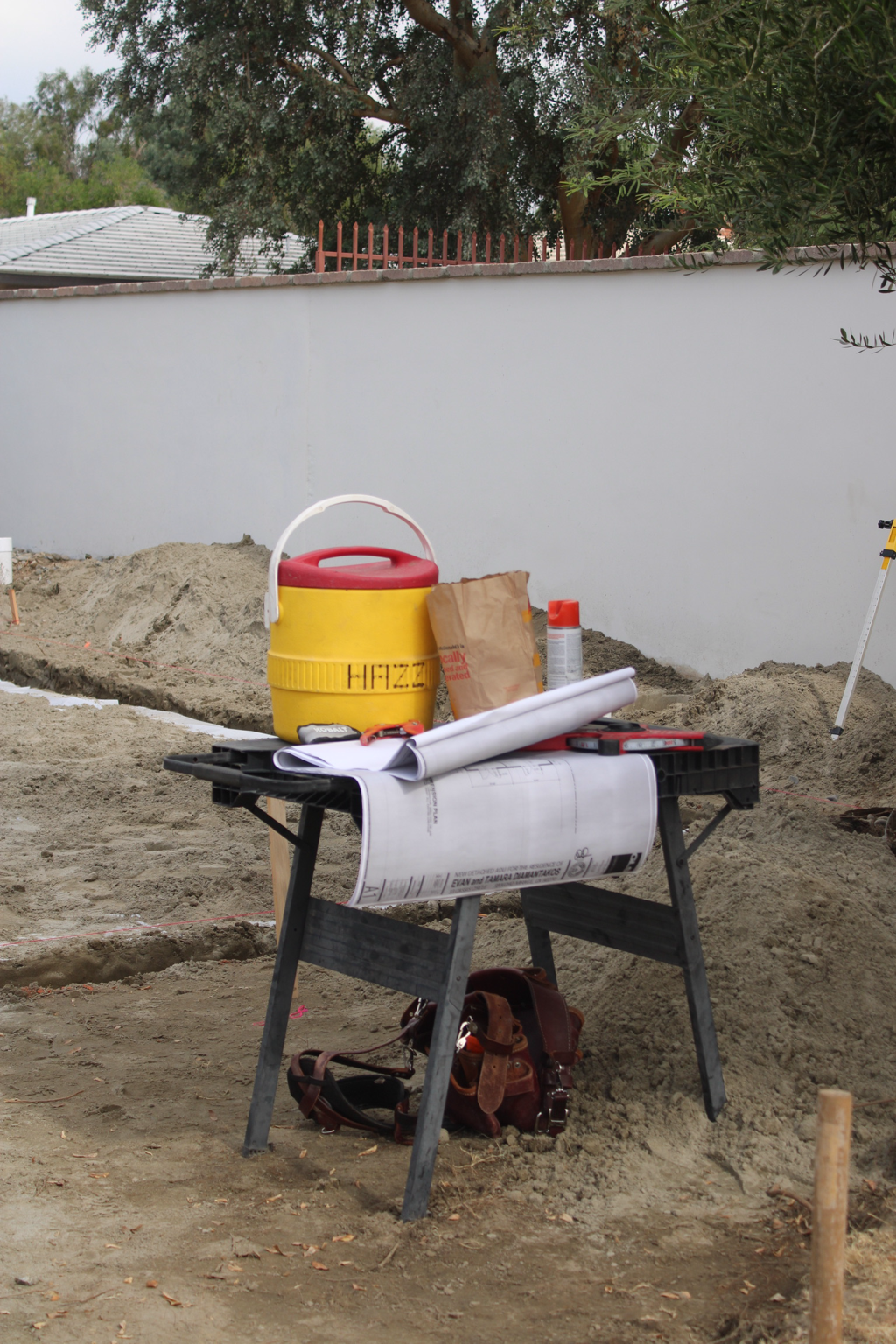 Construction site with a black portable work desk holding blueprints, measuring tools, and a calculator. Behind the desk are a yellow hazard bucket, a paper bag, and a gray spray can. There are trenches in the ground and a white wall with trees and houses in the background.