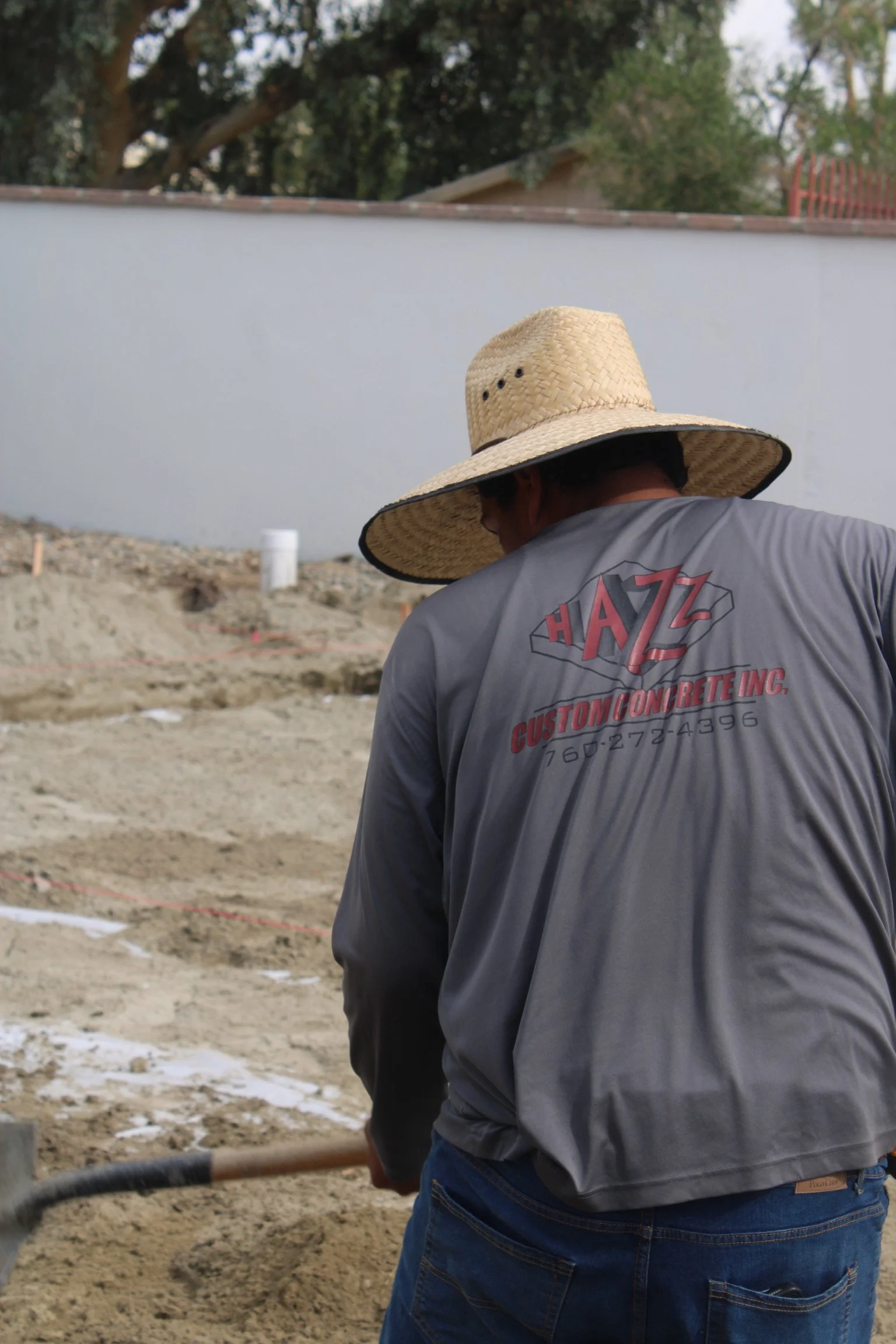 Man wearing a large straw hat and a gray T-shirt with red text working on a construction site with dirt and gravel, some snow, and trees in the background.