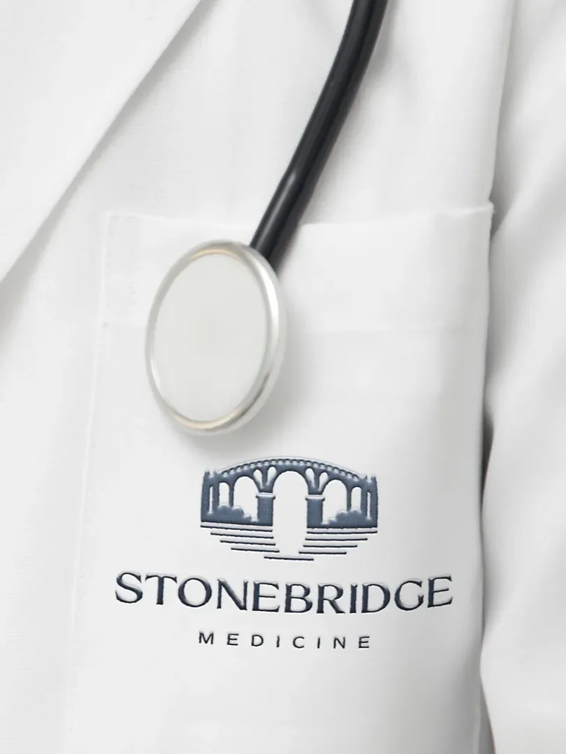 Close-up of a doctor's white coat with the Stonebridge Medicine logo, featuring a bridge graphic, and a black stethoscope draped around the neck.