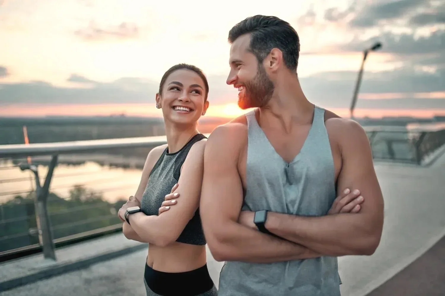 A smiling young woman and man in athletic clothing stand on a bridge at sunset, facing each other with arms crossed.