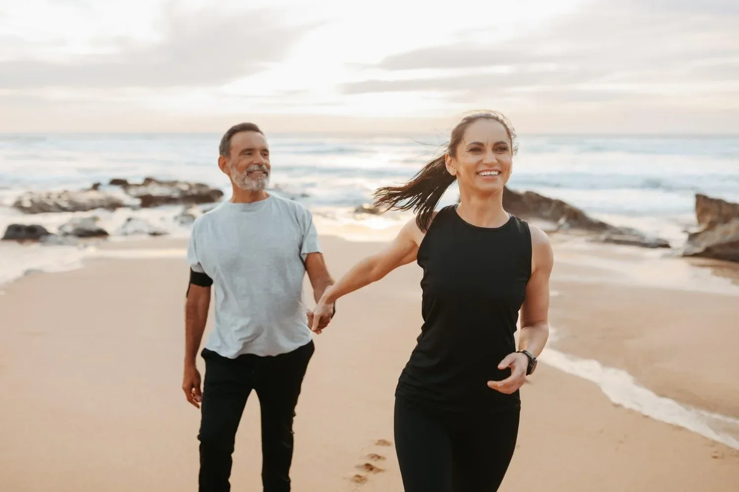A happy woman in black athletic wear running on a beach holding hands with a smiling man in a light gray t-shirt and black pants, during sunset with rocks and ocean in the background.