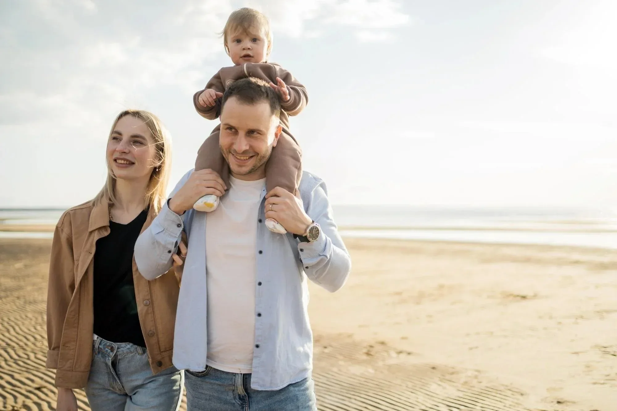 A happy family walking on the beach during daytime, father carrying a young child on his shoulders, mother walking beside them, ocean and cloudy sky in the background.