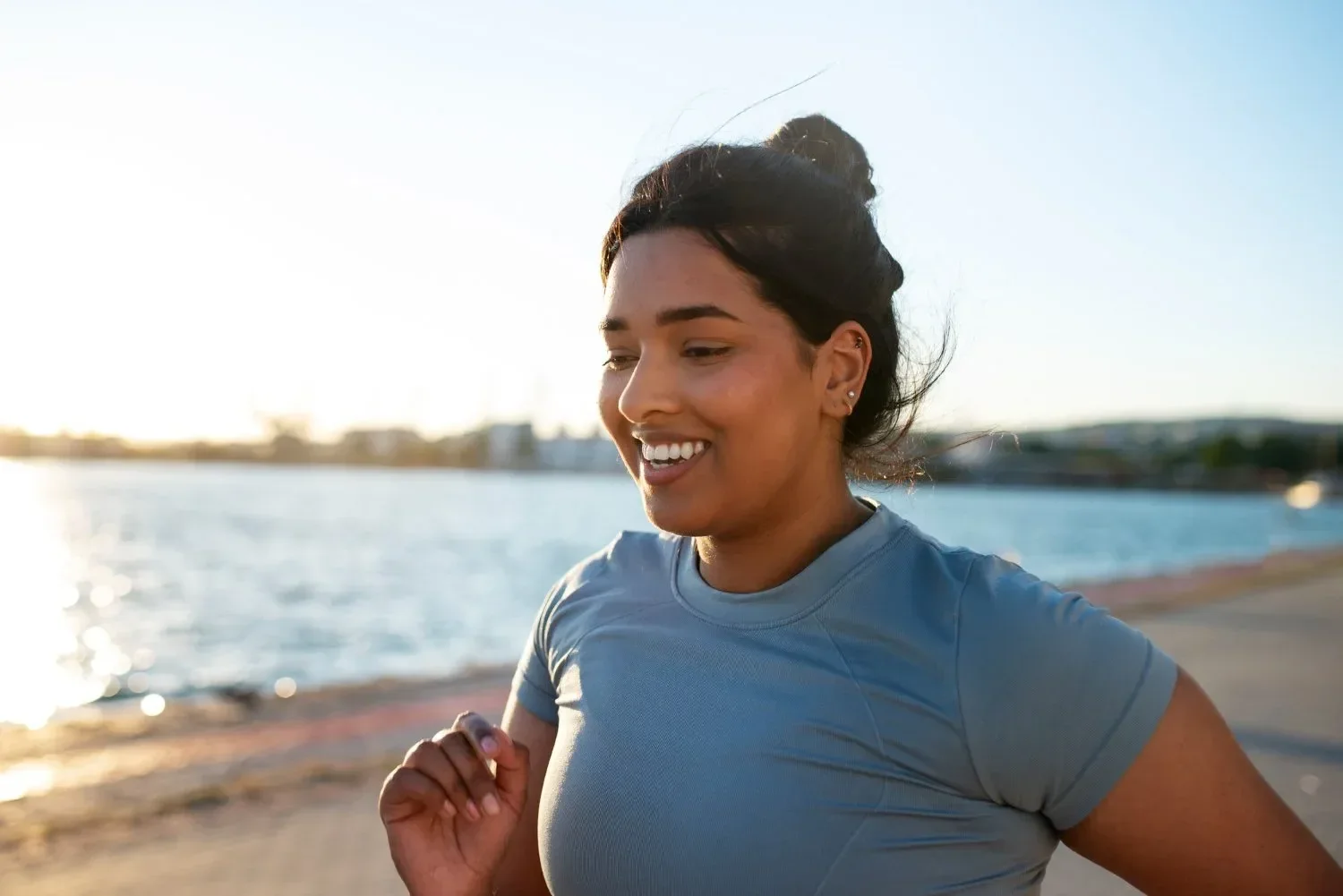 A woman running outdoors along a waterfront during sunset, wearing a light blue athletic shirt, smiling, with her hair in a bun.