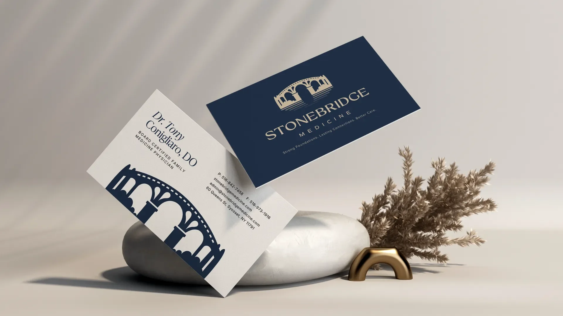 Business cards for Stonebridge Medicine and Dr. Tony Conigliaro, featuring a bridge graphic, placed on a smooth stone with dried beige pampas grass and a small gold object, set against a neutral background.