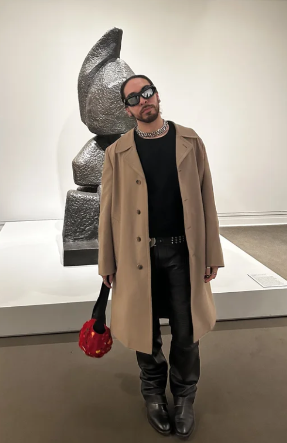 A person with long hair, beard, wearing sunglasses, a beige coat, and black pants, standing in an art gallery in front of a metallic abstract sculpture.