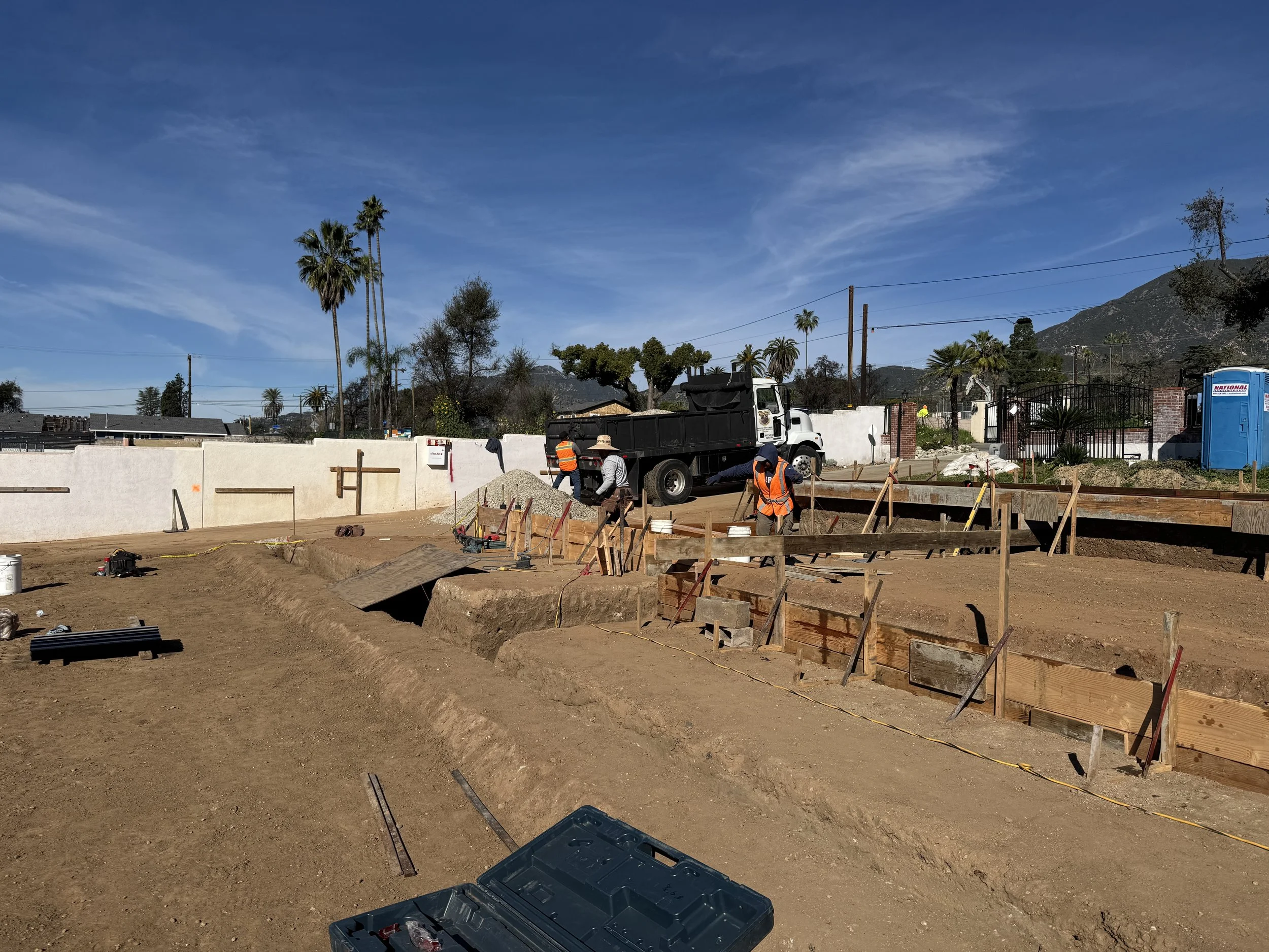 Construction site with workers installing concrete forms, dirt ground, truck, and blue portable toilet under a clear blue sky with trees and mountains in the background.