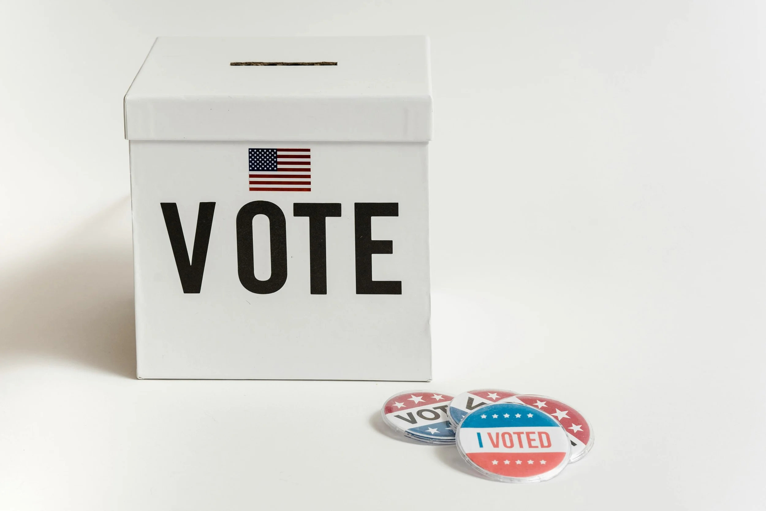 A white ballot box with a slot on top, labeled 'VOTE' with an American flag above, and three 'I VOTED' pins on a white surface.
