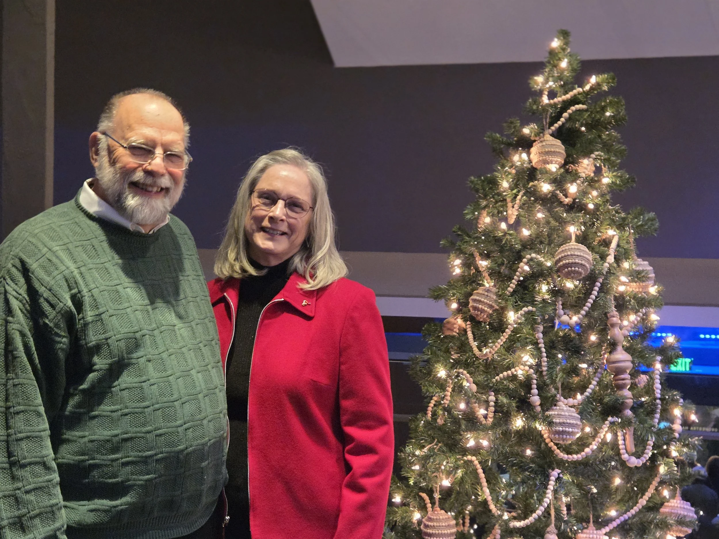 A smiling Mark Jantzer and his wife standing next to a decorated Christmas tree with lights and ornaments.