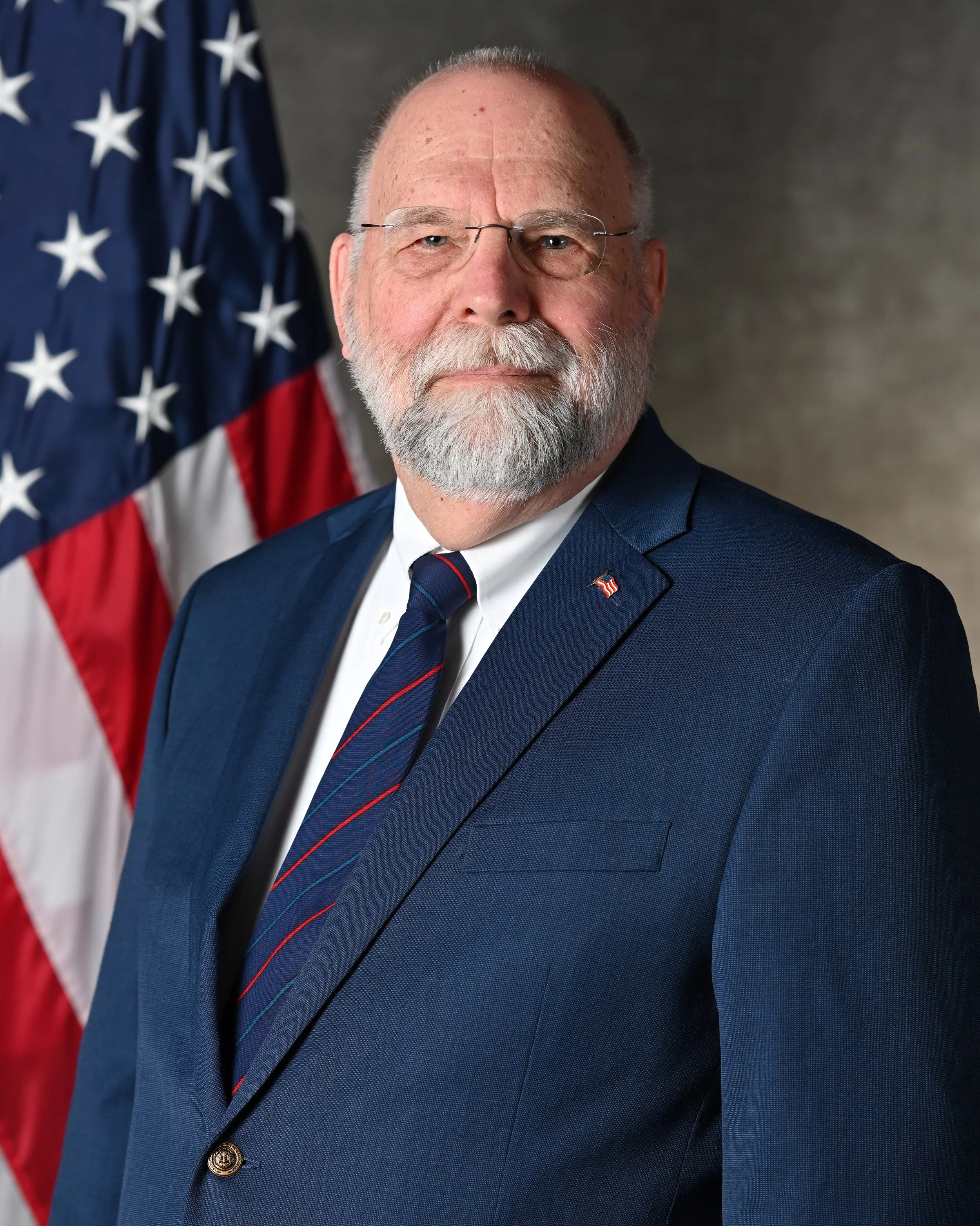 Portrait of Mark Jantzer, wearing a navy suit, white shirt, and a blue tie with red stripes, standing in front of a U.S. flag.