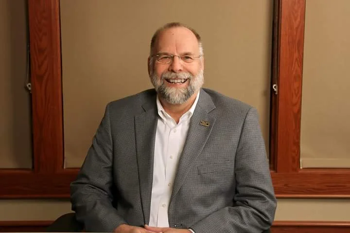 A smiling middle-aged man with a beard and glasses, wearing a gray blazer and white shirt, sitting in front of a wooden-framed window with beige blinds.
