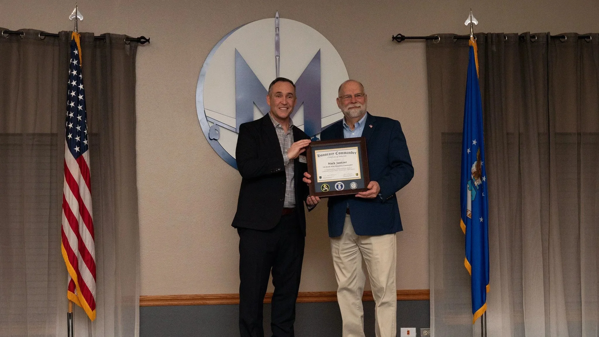 Two men in suits holding a framed certificate and smiling at the camera. One man has short dark hair and the other has gray hair and a beard. They are standing in front of a large logo with the letter 'M' and flags of the United States and Missouri behind them.