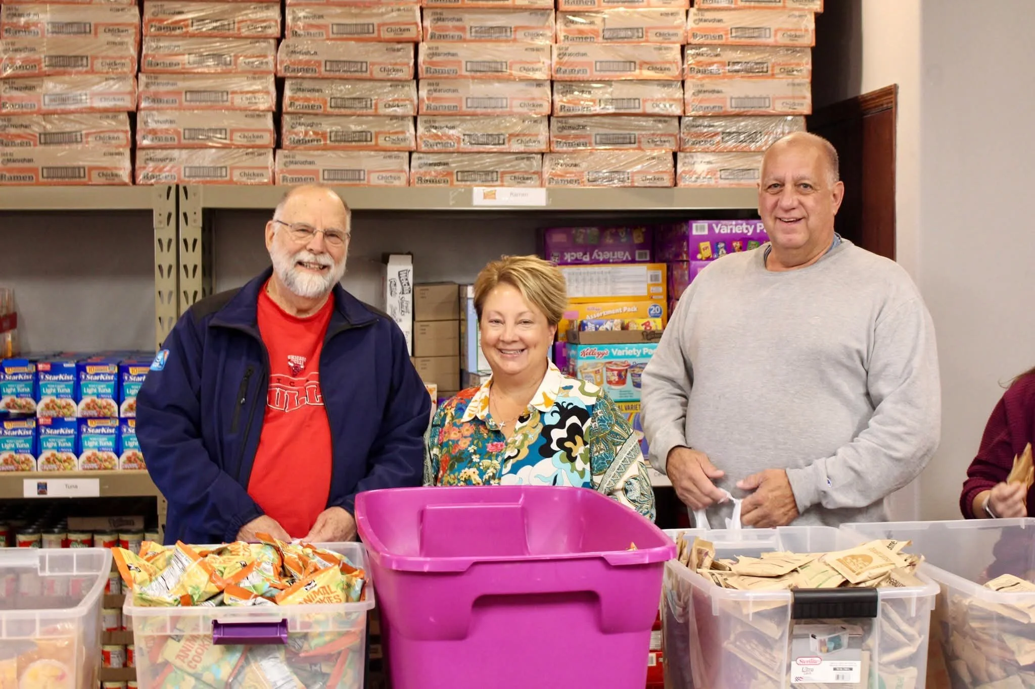Mark Jantzer supporting local nonprofits, standing behind tables with various food items in plastic bins, smiling in a warehouse or store aisle with shelves stocked with boxed food products.
