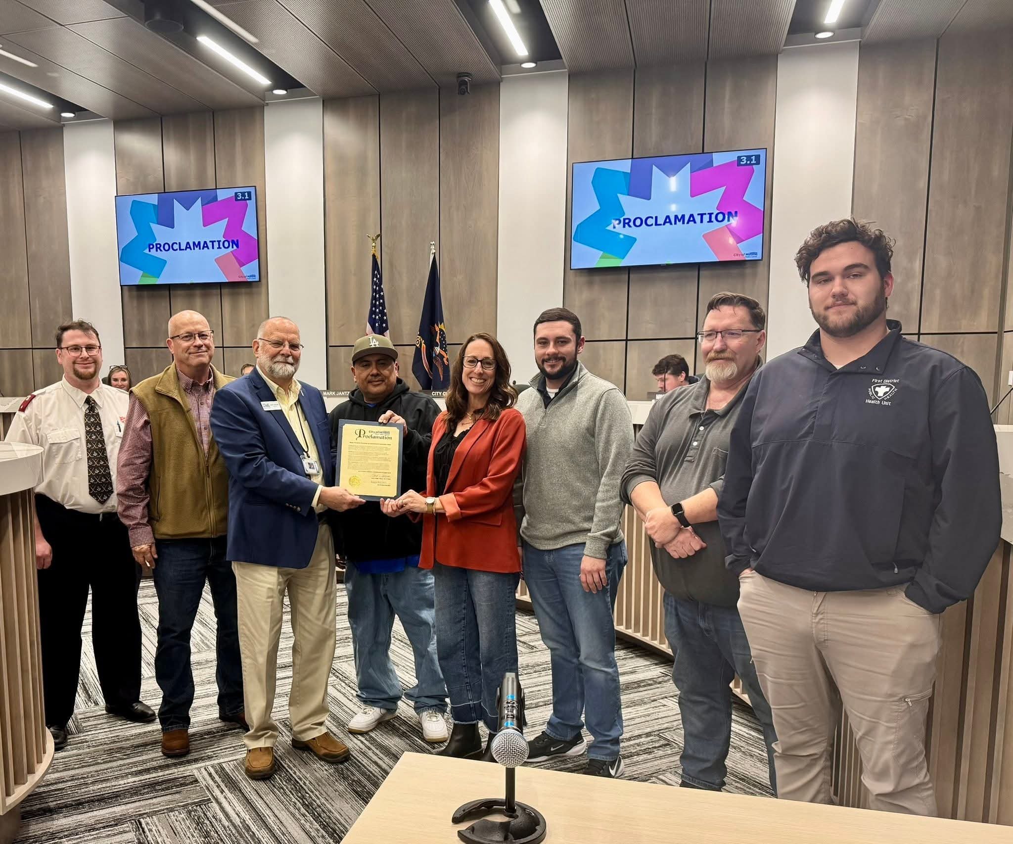 A group of nine people standing in a conference room, with two of them holding a proclamation document. The background shows two screens displaying the word 'Proclamation' and flags behind them.