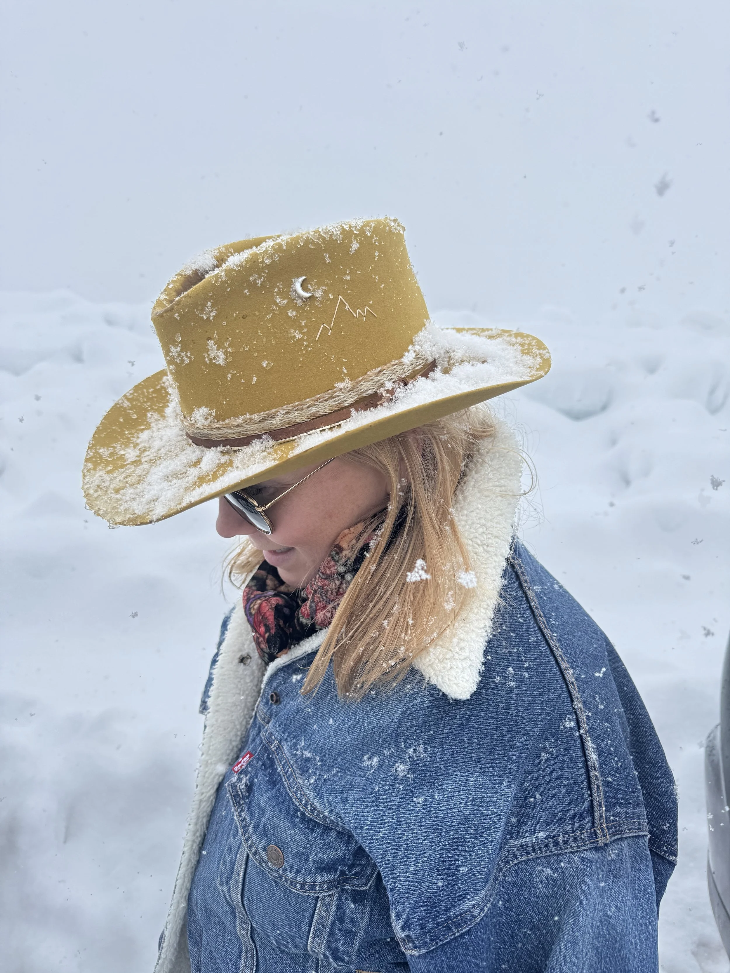 Close-up of a woman wearing a yellow felt cowboy hat with snow on it, standing outdoors in a snowy landscape.