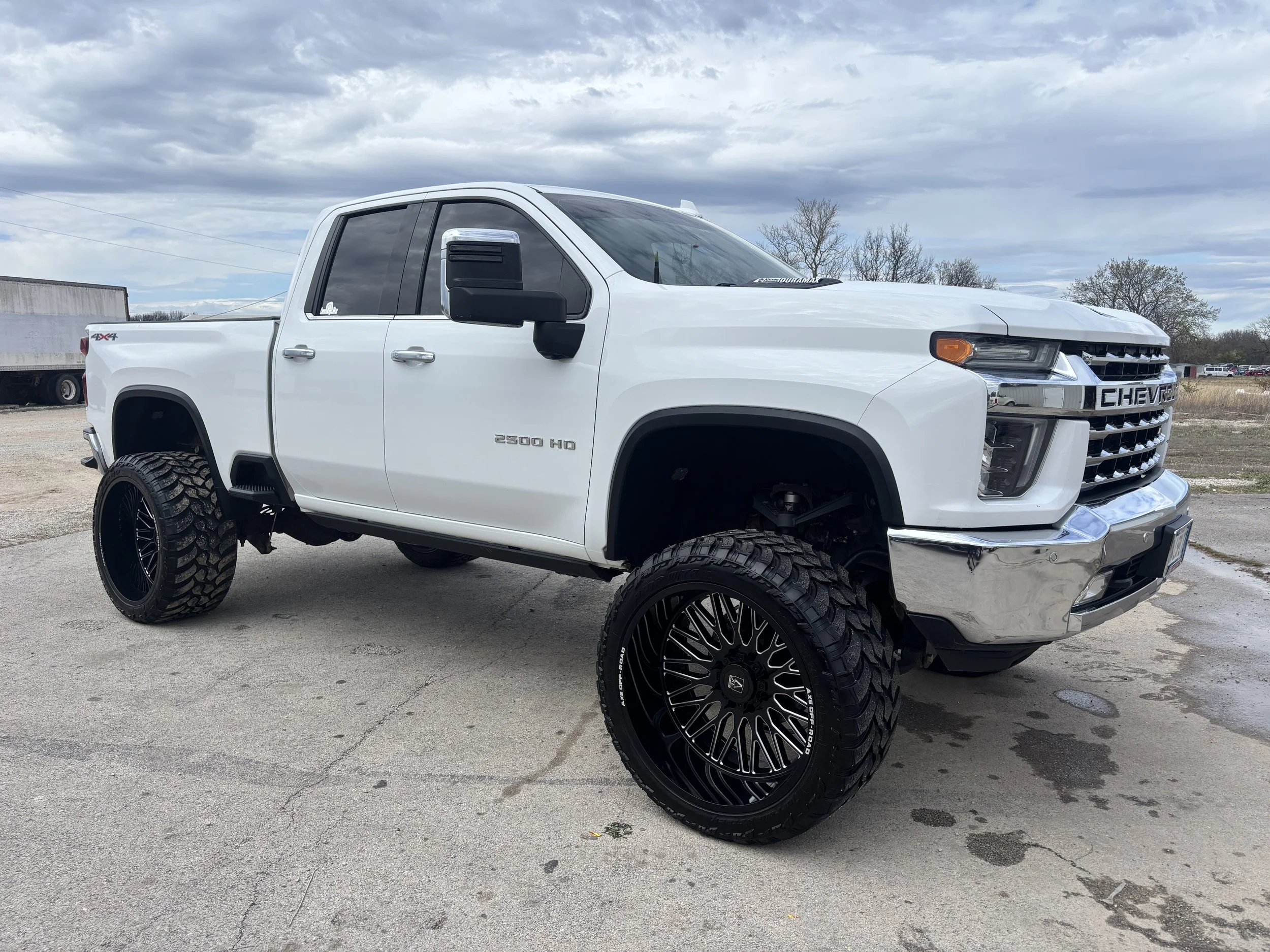 White Chevrolet Silverado 2500 HD pickup truck with large black wheels and off-road tires parked outdoors on concrete, under a cloudy sky.
