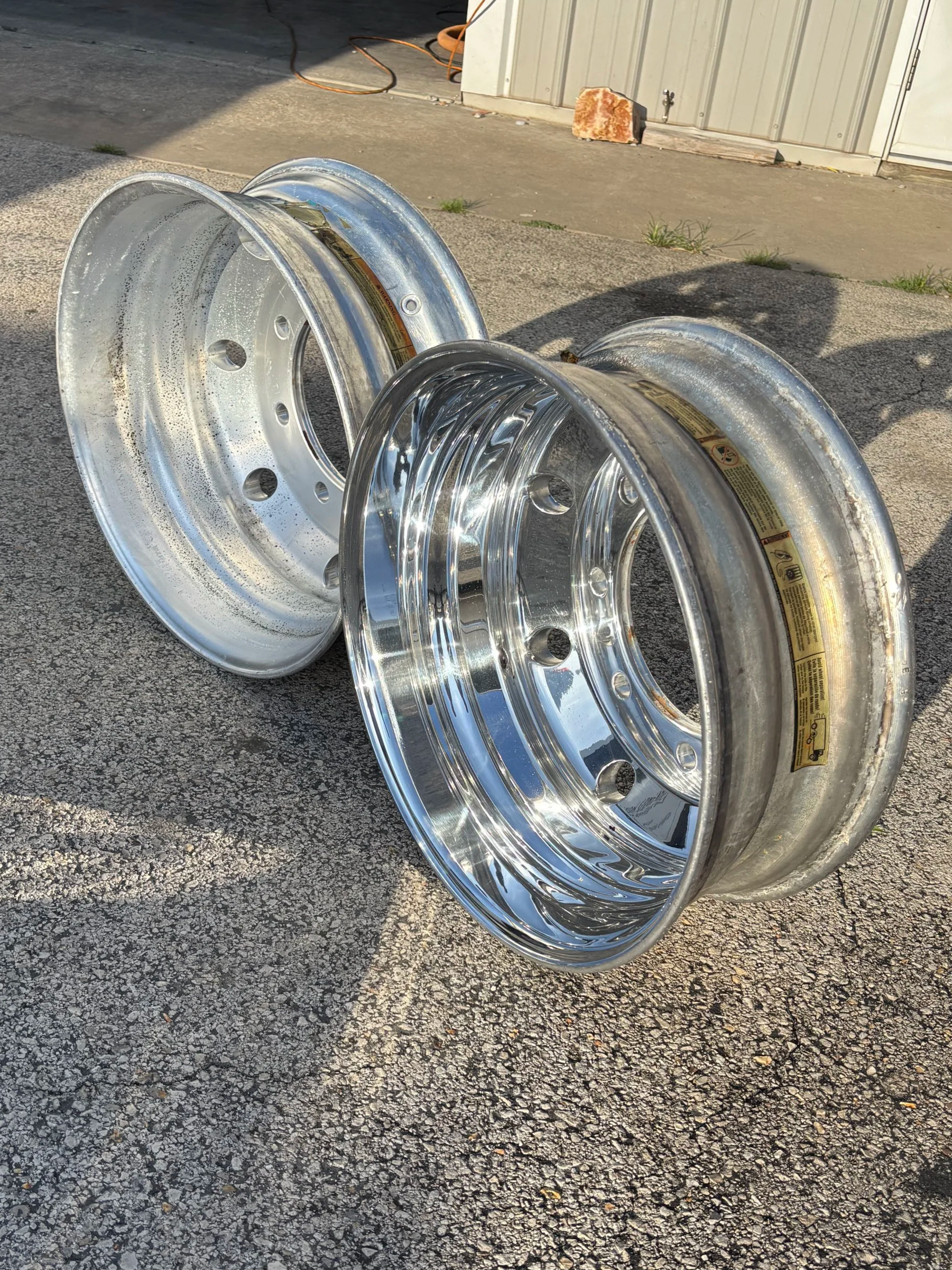 Two shiny, chrome-plated metal rims lying on an asphalt surface outdoors, with a shed and some small grass patches in the background.