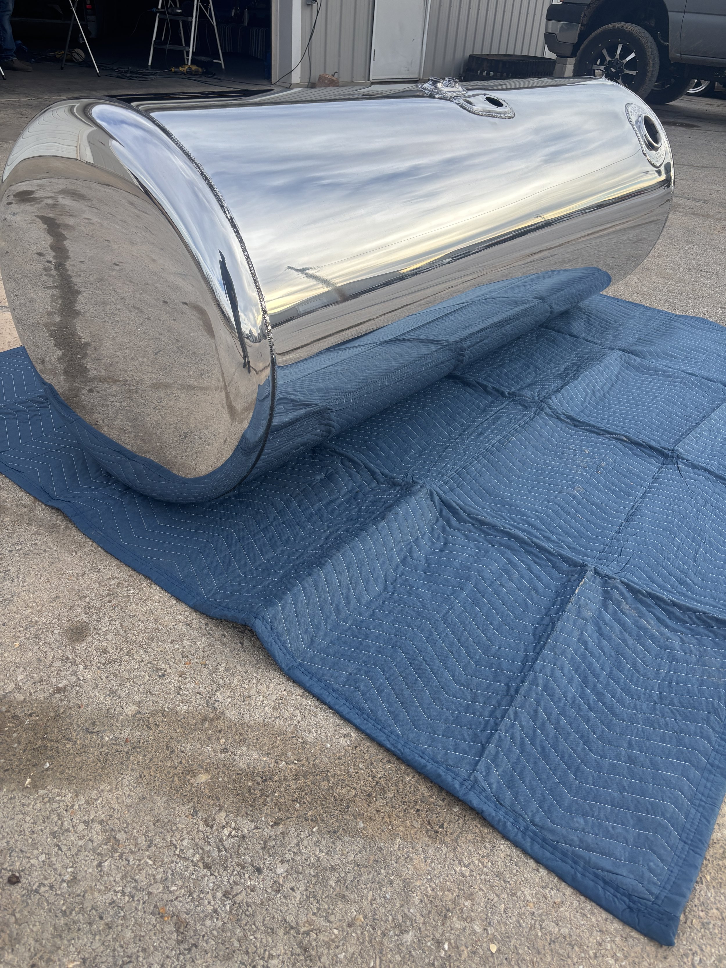 A shiny, metallic cylindrical tank resting on a blue padded blanket outdoors, with a garage and a pickup truck visible in the background.