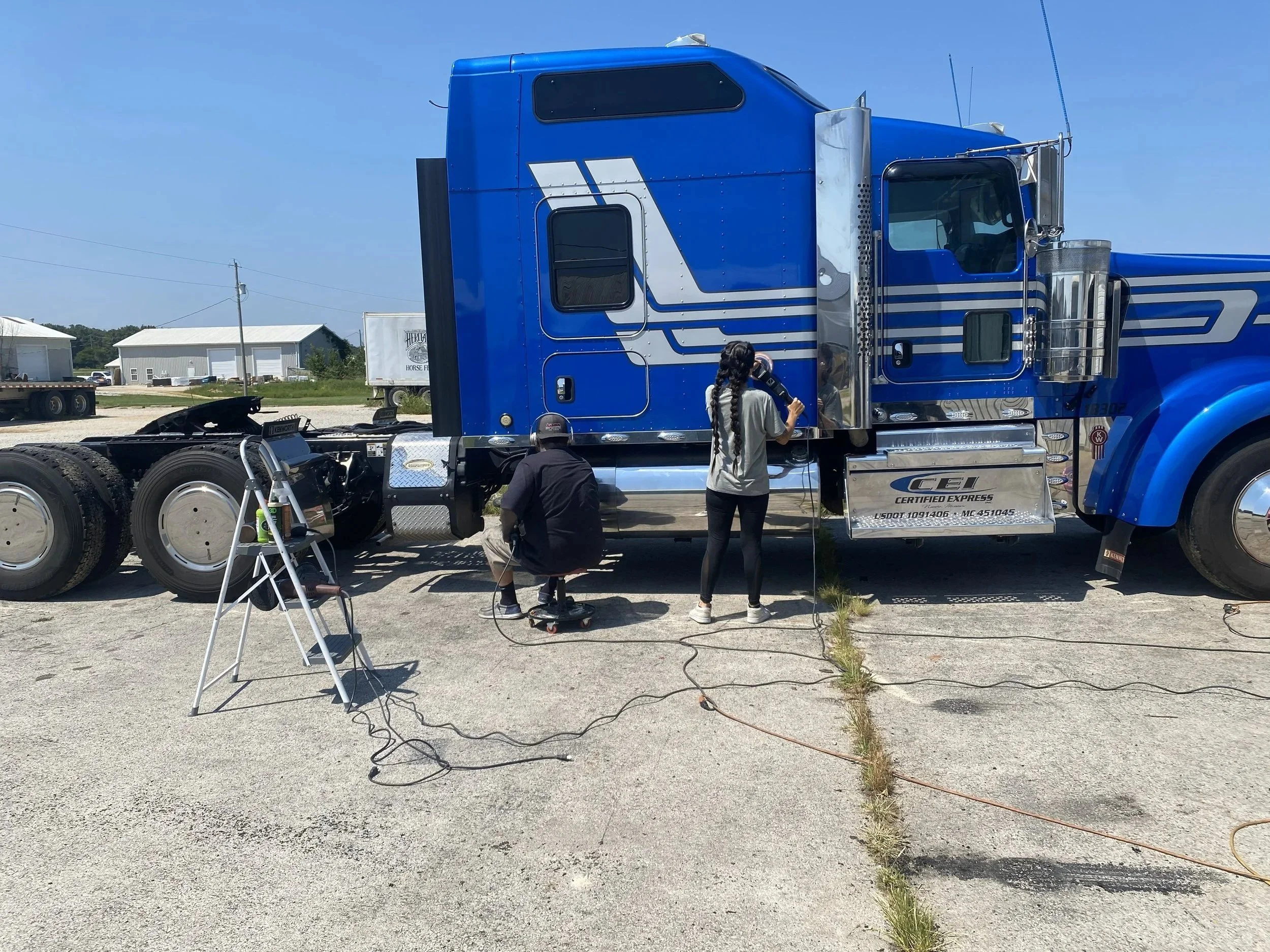 Two people working on or inspecting a large blue semi-truck parked outside on a sunny day.