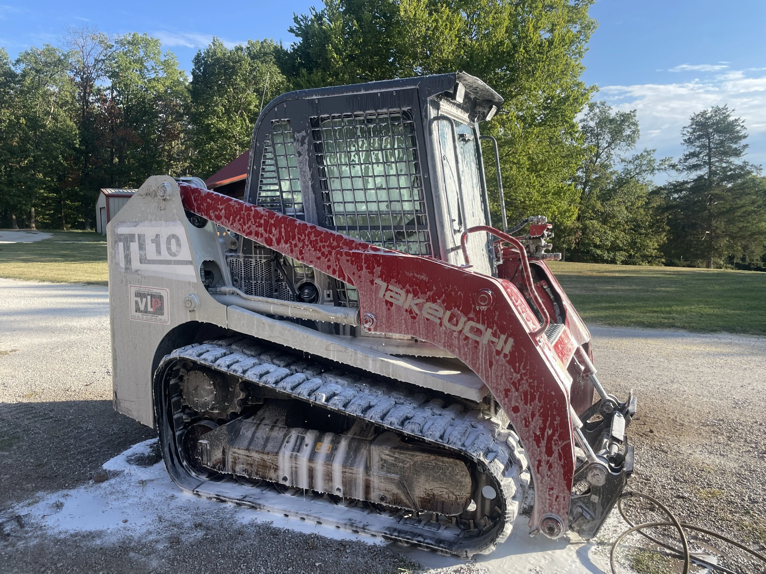 A red and white skid-steer loader with foam on the tracks, parked on gravel outside with trees and a blue sky in the background.