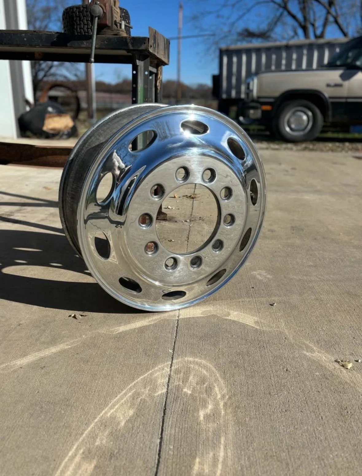 A shiny metal wheel rim placed on a concrete surface outdoors, with vehicles and equipment in the background.