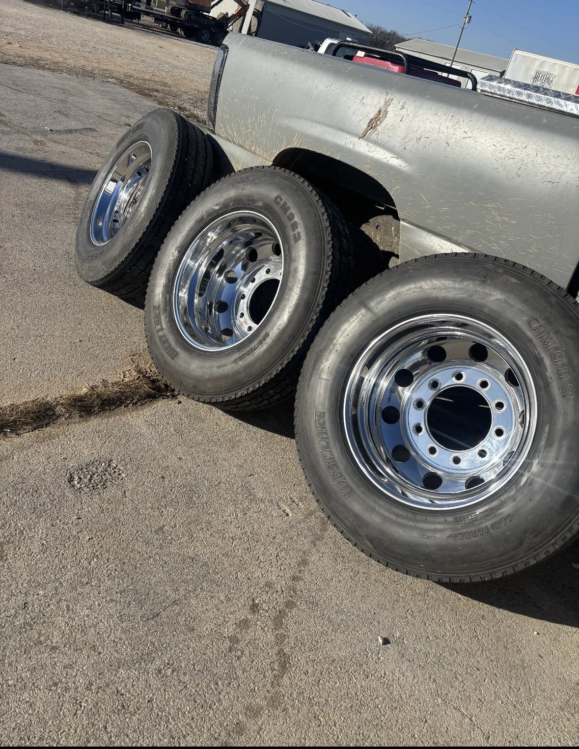 Three large truck tires with chrome rims leaning against the side of a silver pickup truck, situated on a gravel and asphalt surface outside.