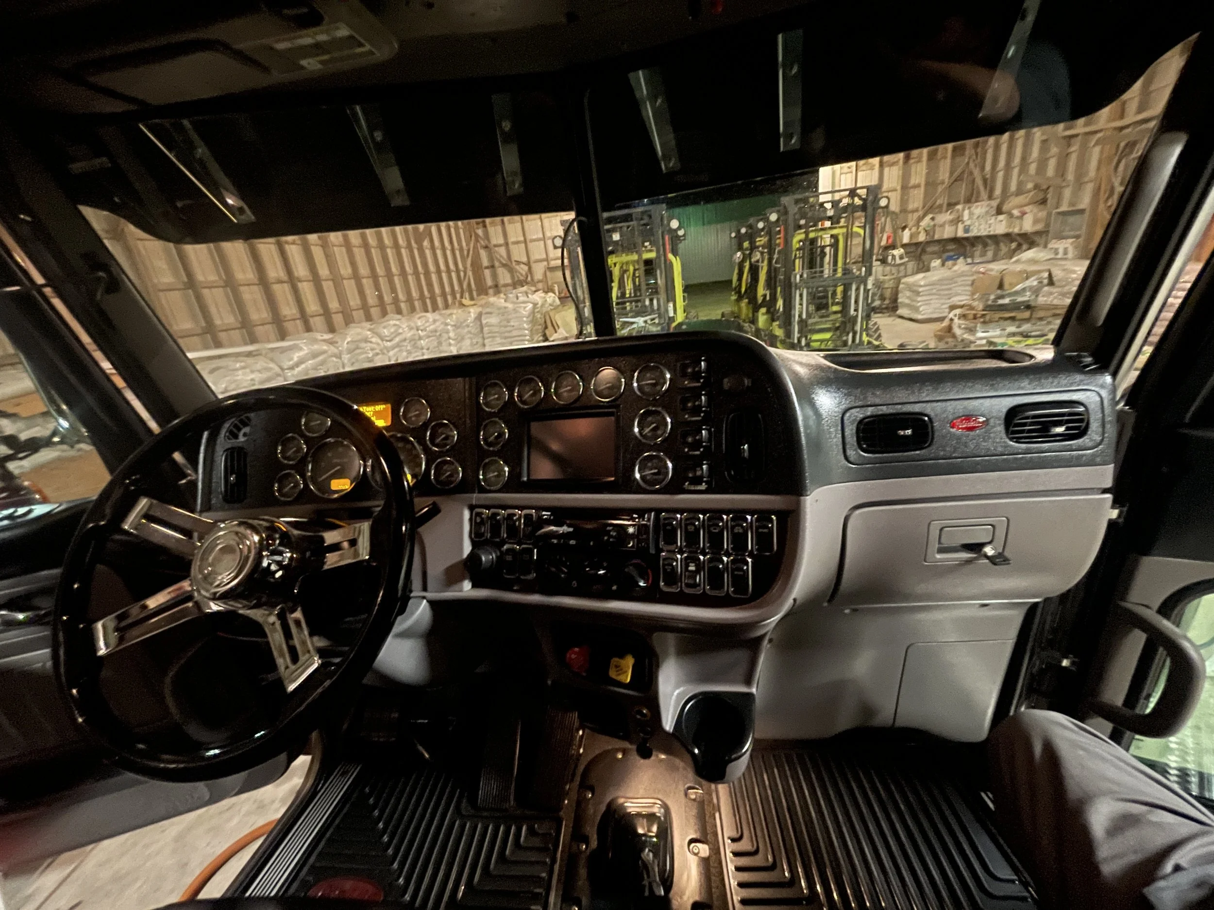 Interior of a semi-truck cab showing dashboard, steering wheel, and controls, with forklifts and warehouse materials visible through the windshield.