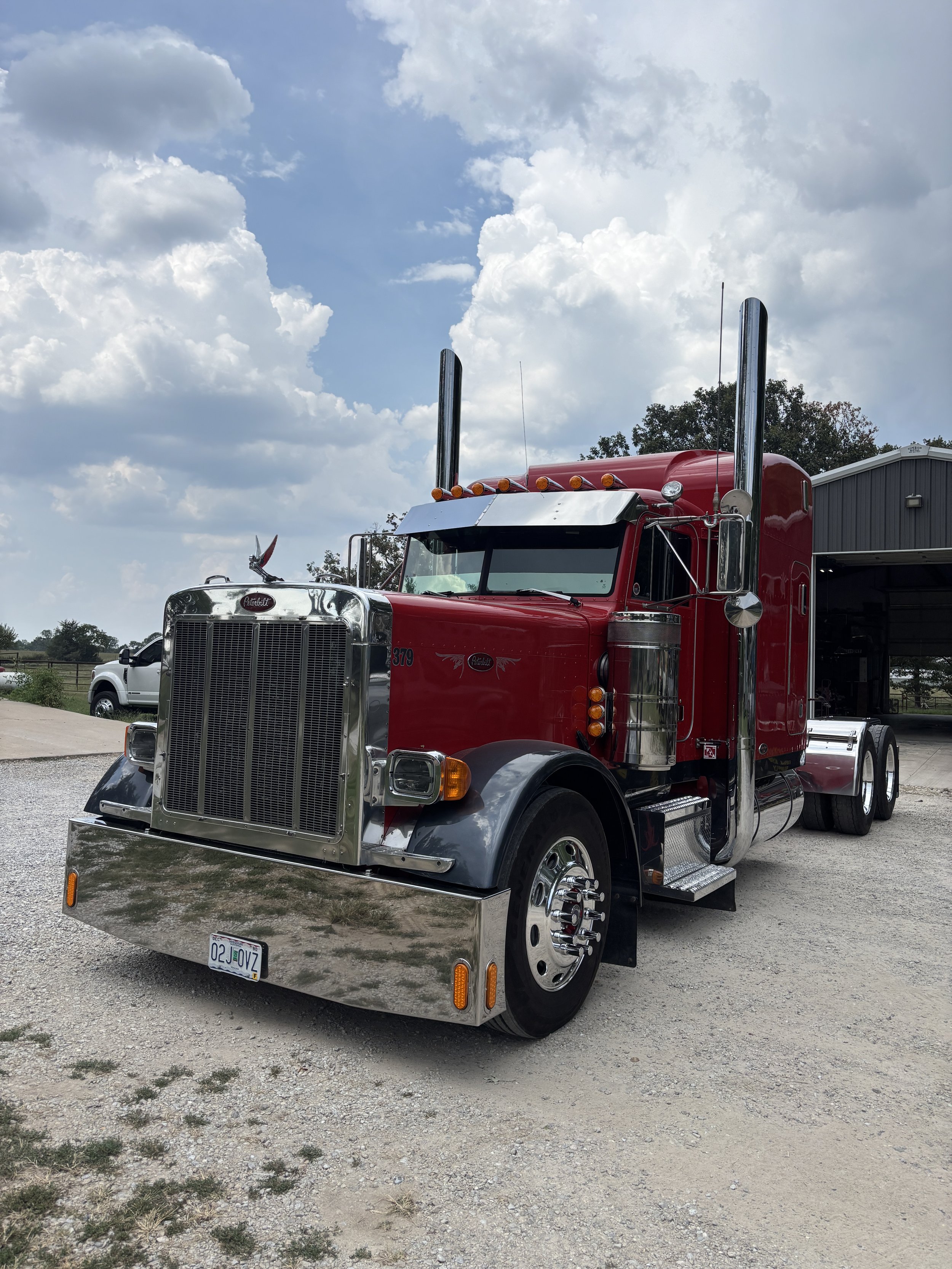 Red semi-truck parked on gravel with a gray warehouse and white vehicle in the background under a partly cloudy sky.