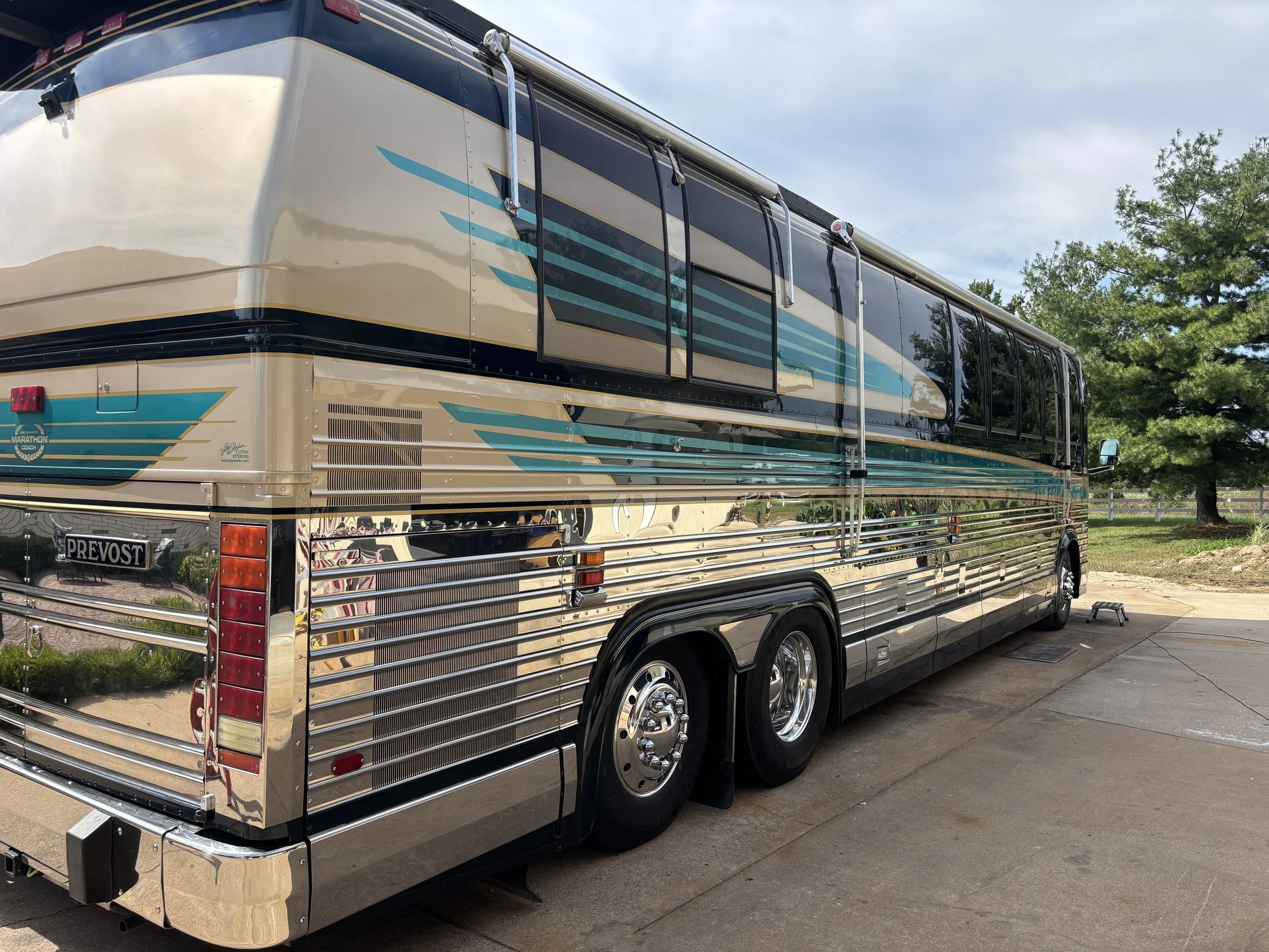 A shiny, vintage Prevost bus with a metallic exterior parked outdoors, with trees and a partly cloudy sky in the background.