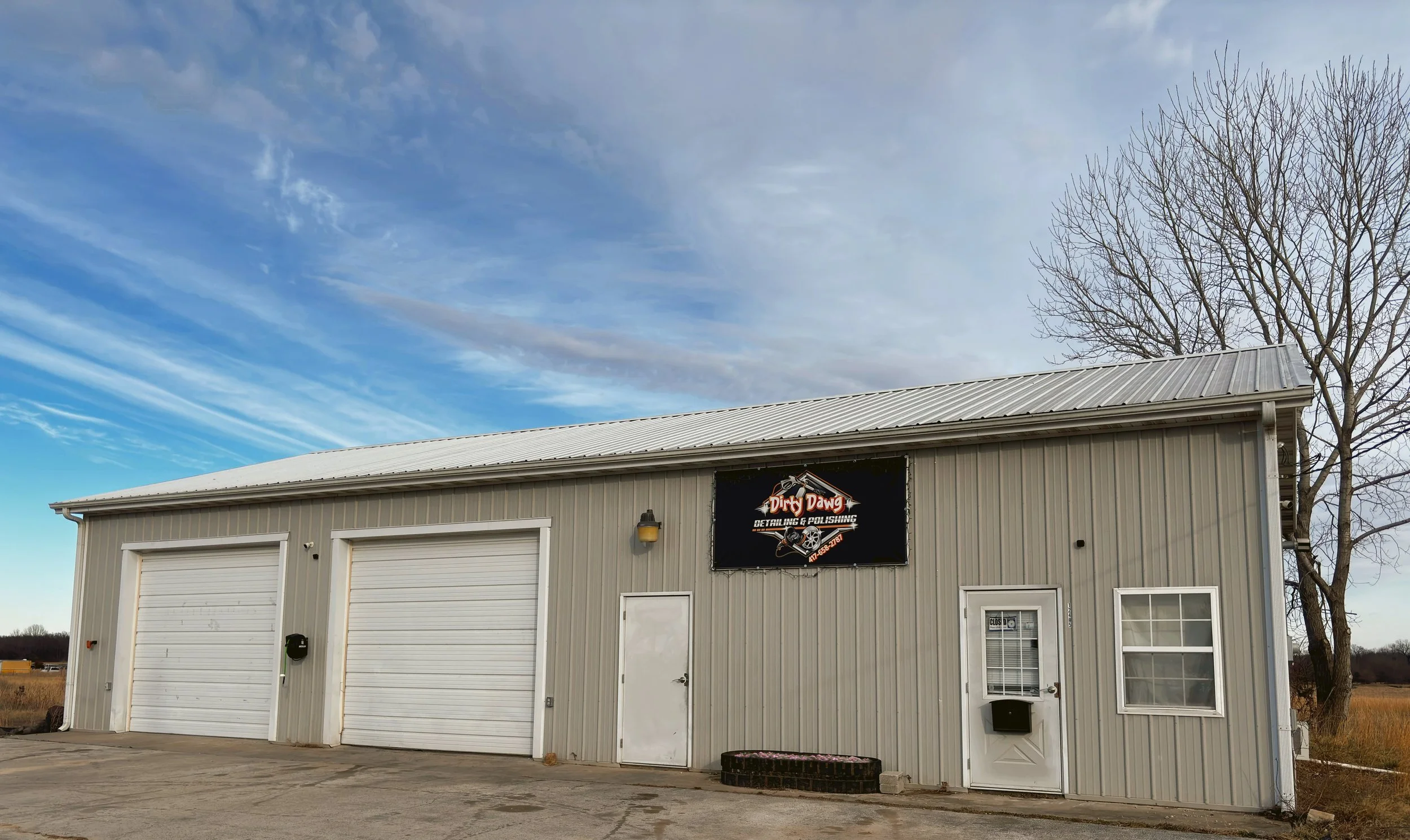 Metal building with two garage doors, a side door, and a sign reading "Dirty Dawg Detailing & Polishing" on a partly cloudy sky background.