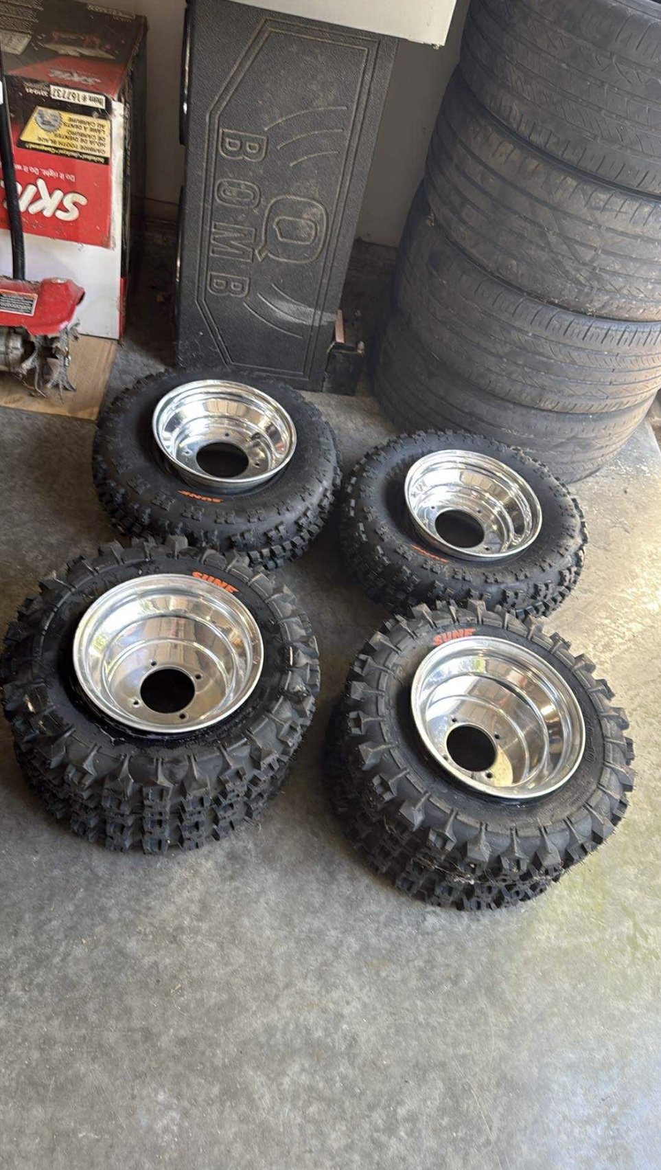 Four off-road tires with shiny metal rims on a concrete floor in a garage or workshop setting.