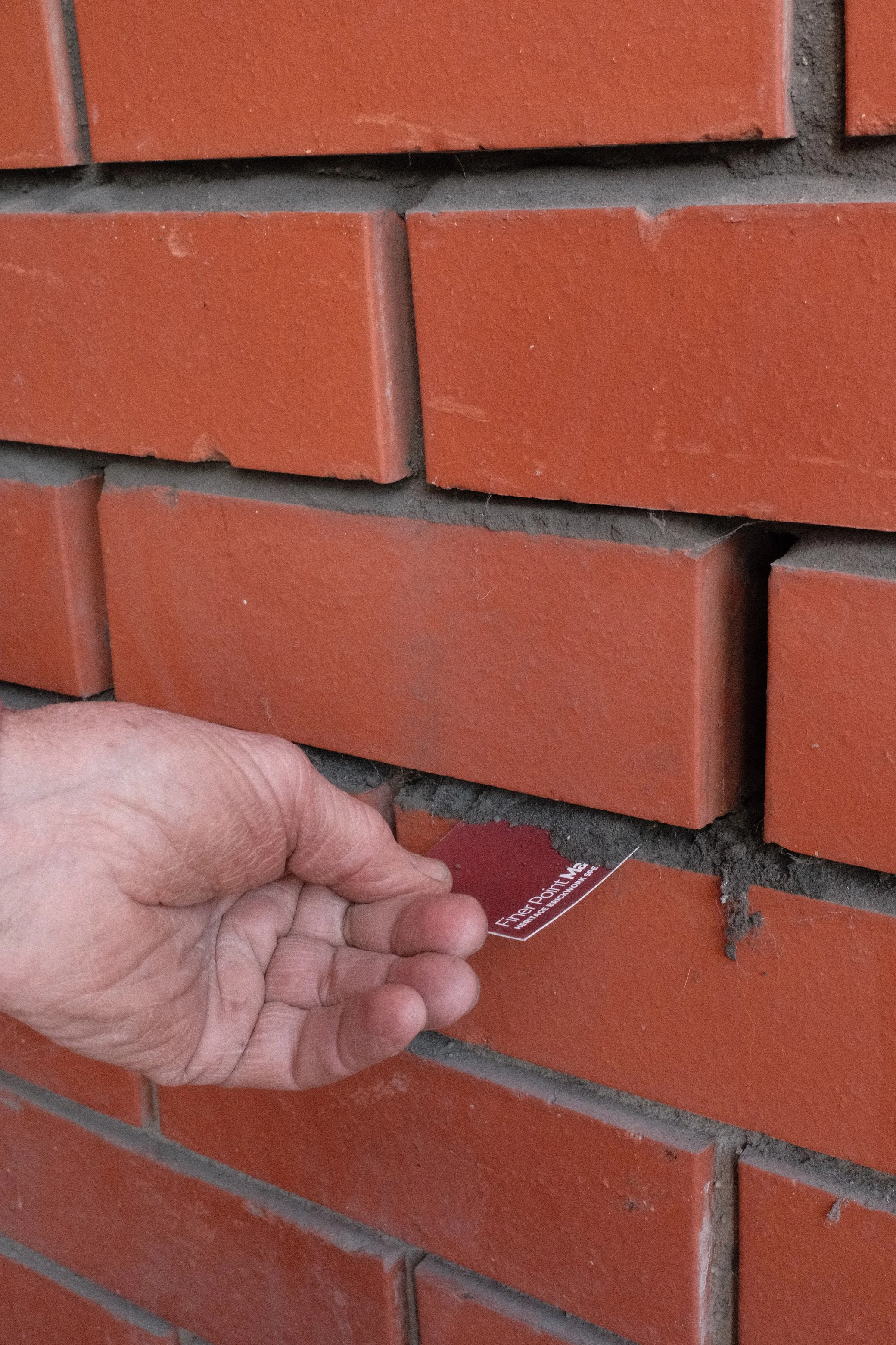 Close-up of a person's hand inserting a test marker into mortar joint of red brick wall during building inspection.