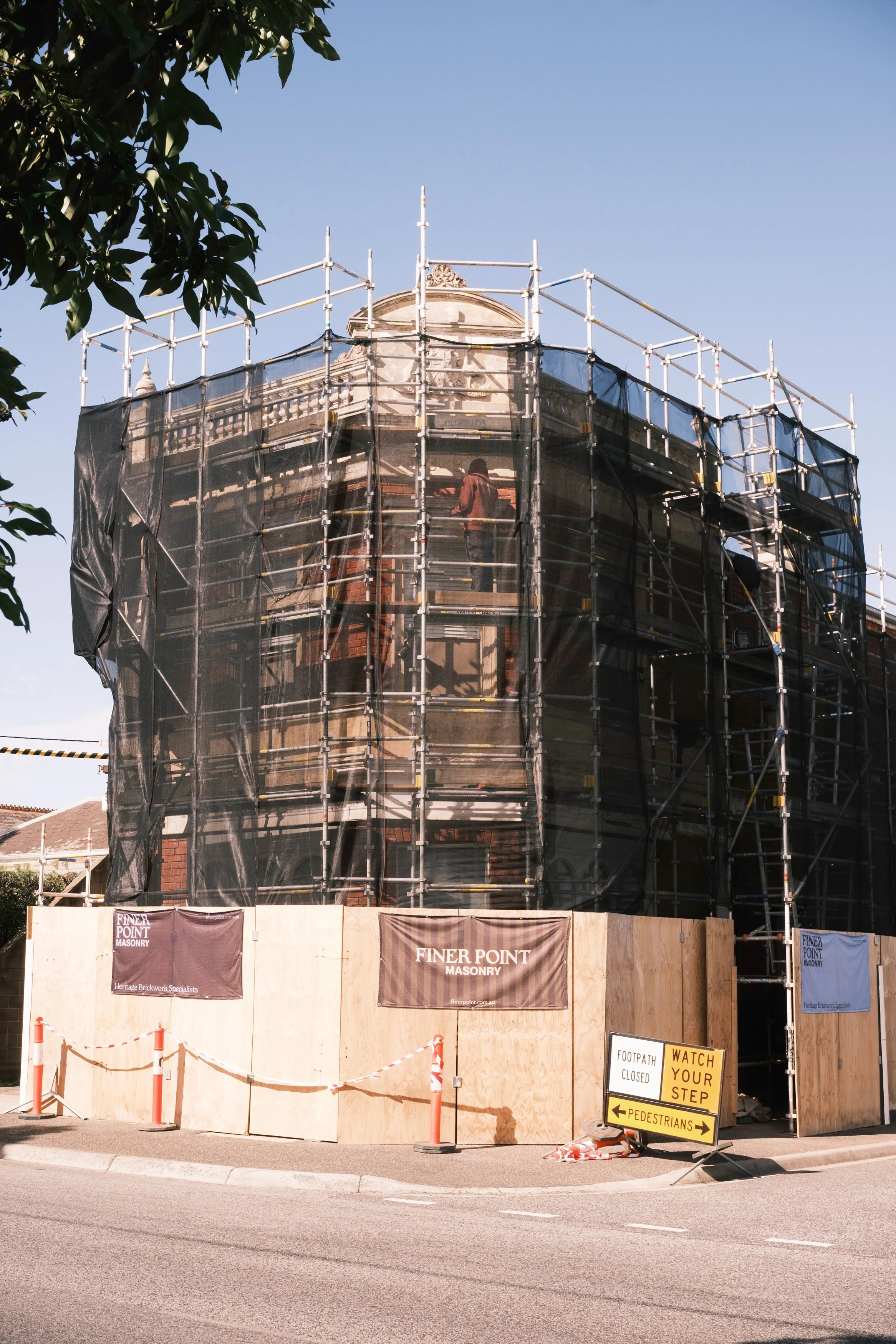 A building under renovation covered with black mesh and wooden fencing, with scaffolding around it, and a sign in the foreground that says 'Footpath Closed' and 'Watch Your Step'.