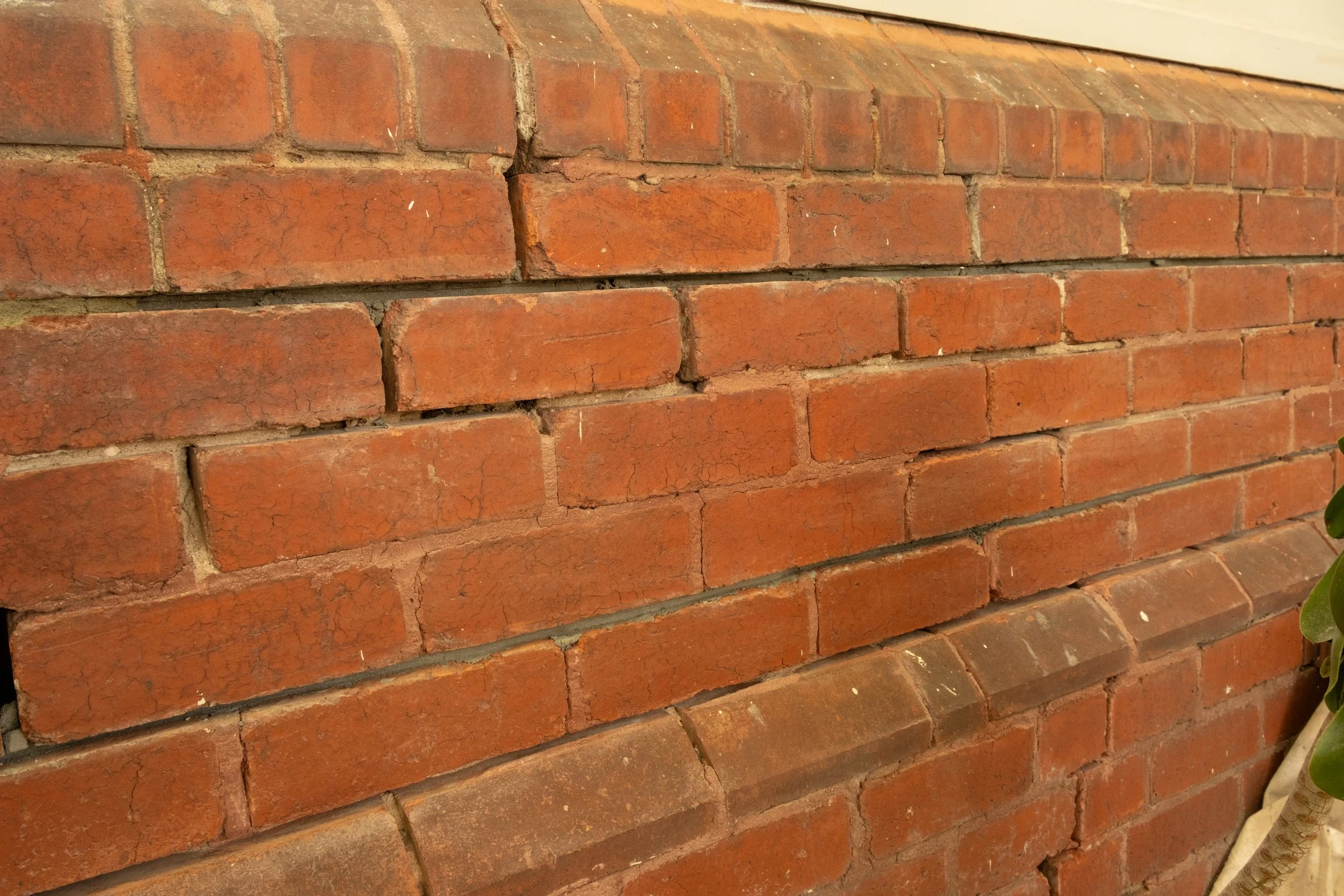 Close-up of a red brick wall with some bricks cracked and uneven, and a small part of a leaf visible on the bottom right corner.