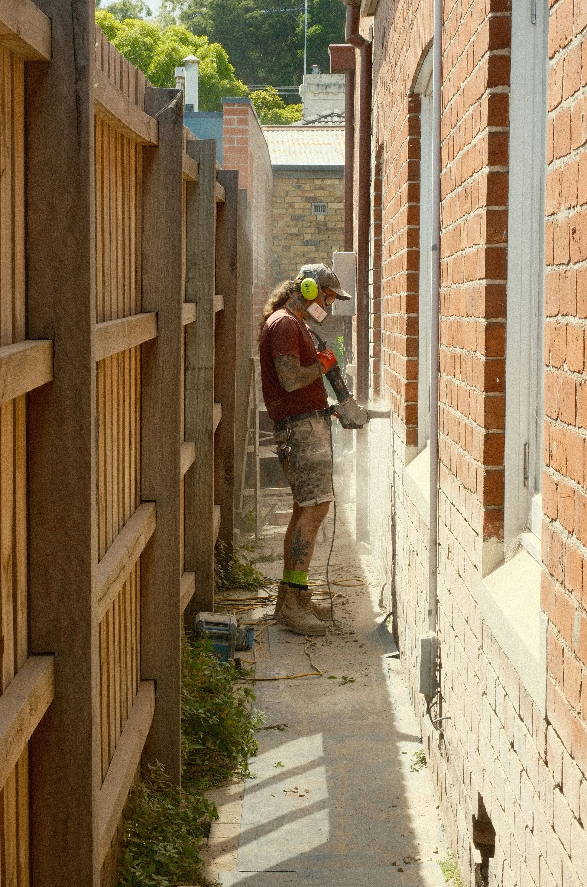 Man working on brick wall with power tool in a narrow alley between a brick house and wooden fence on a sunny day.