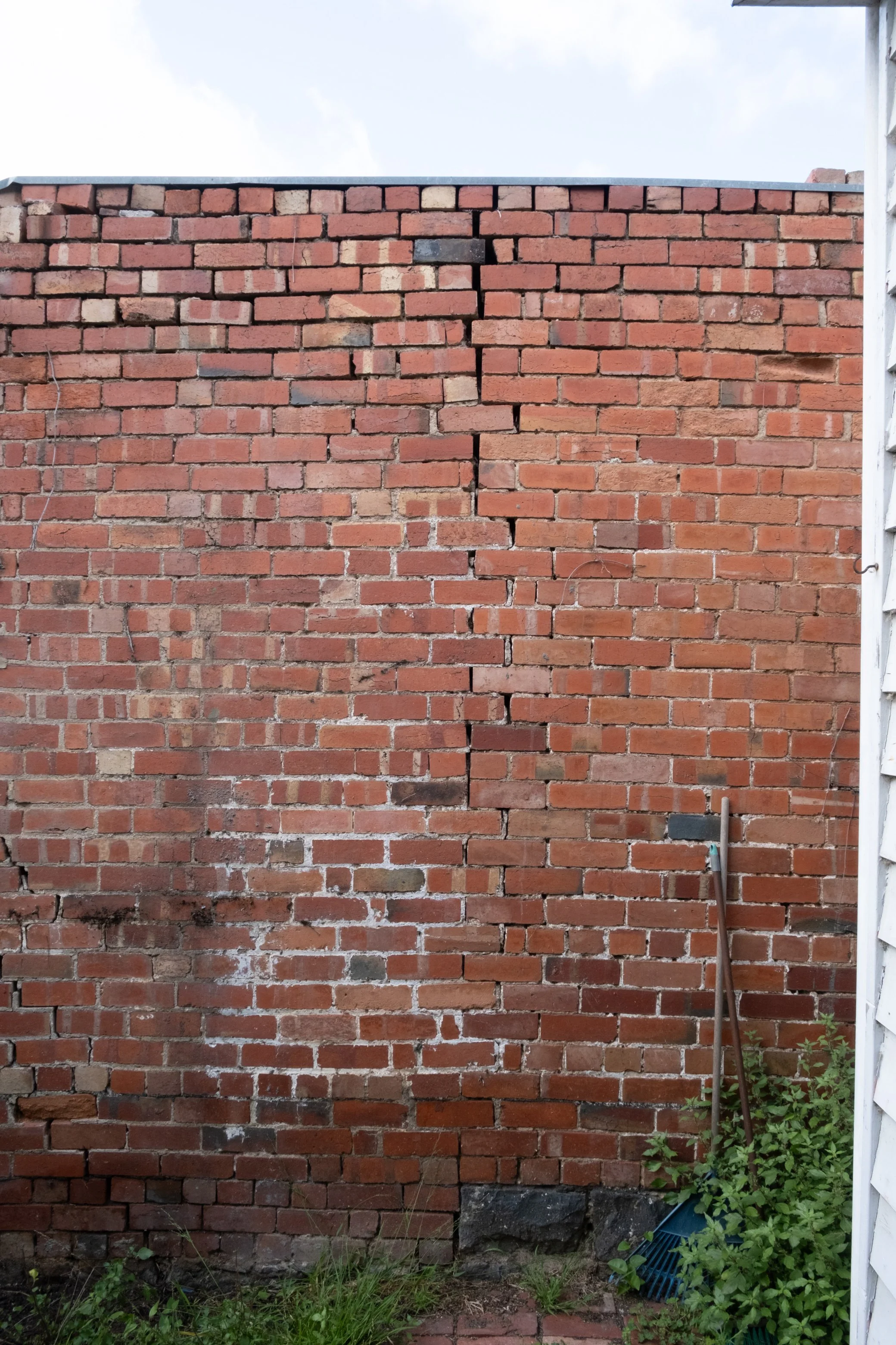 A brick wall with a visible crack running vertically down the middle, surrounded by some grass, dirt, and plants at the base, and a small blue rake leaning against the wall on the right side.